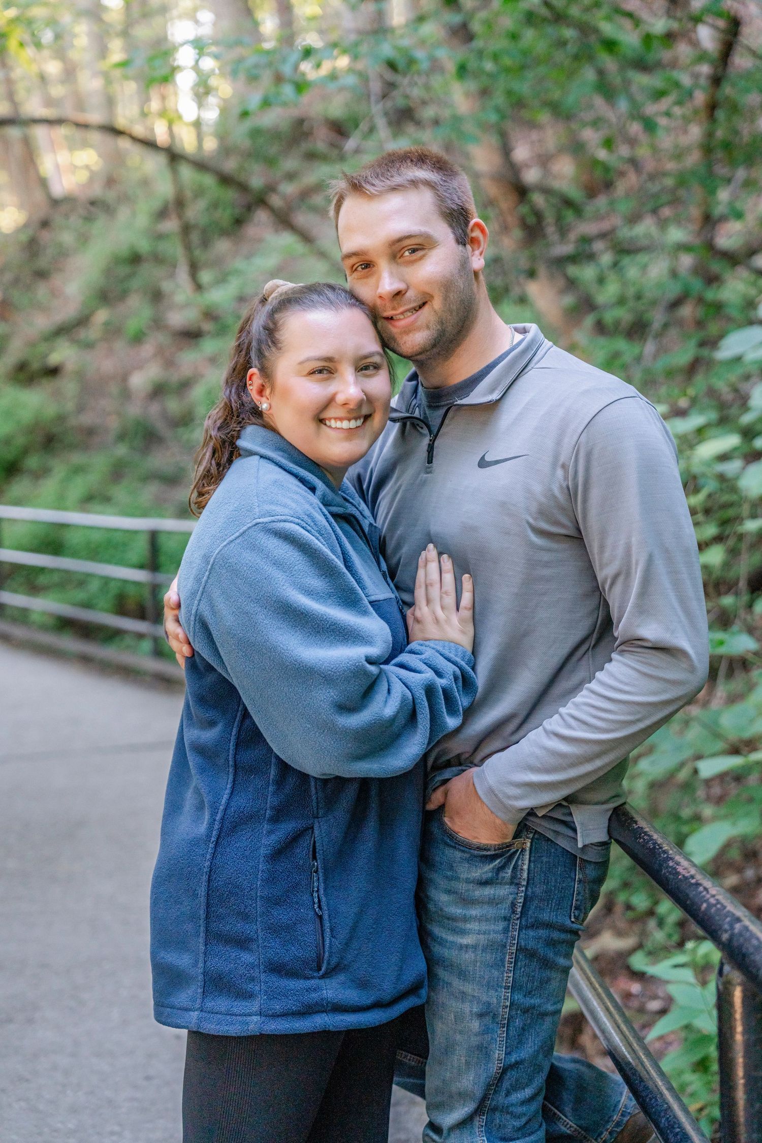 Two people embrace and smile while standing on a nature path surrounded by green foliage.