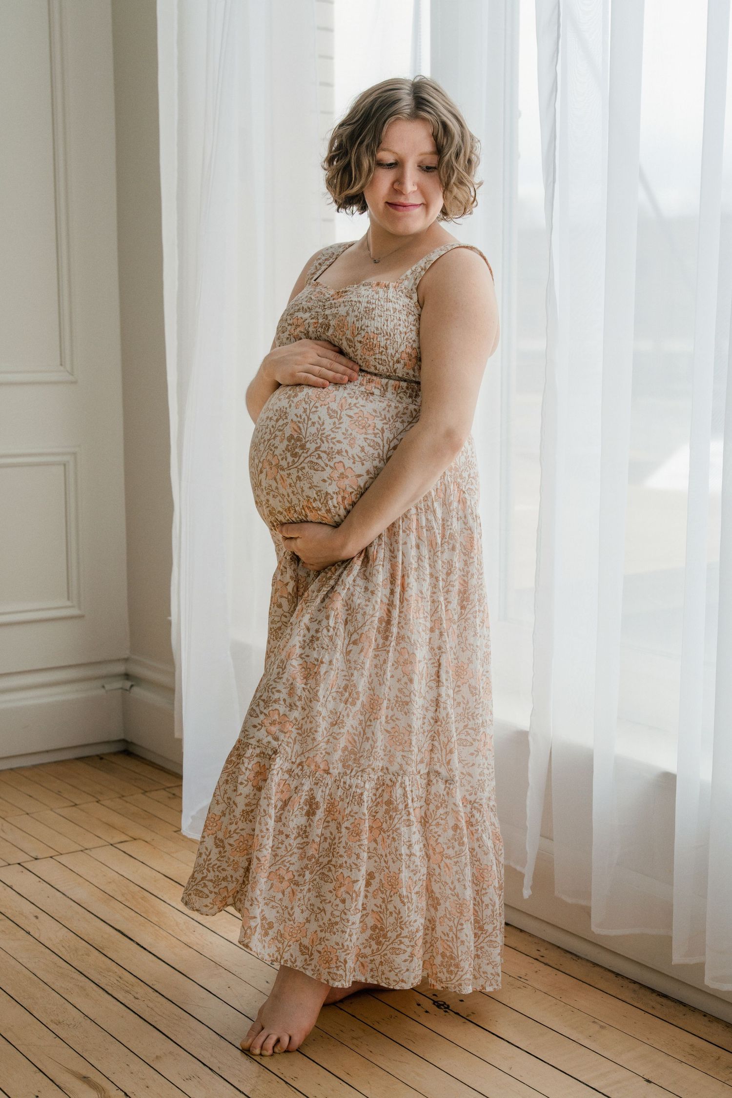 An expectant mother wearing a floral maxi dress poses barefoot by sheer curtains in a room with hardwood floors.