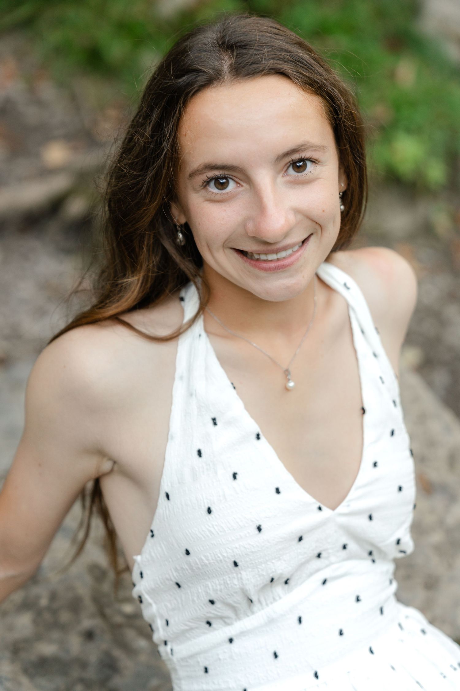A person wearing a white sleeveless top with small dark patterns poses outdoors against a natural background.