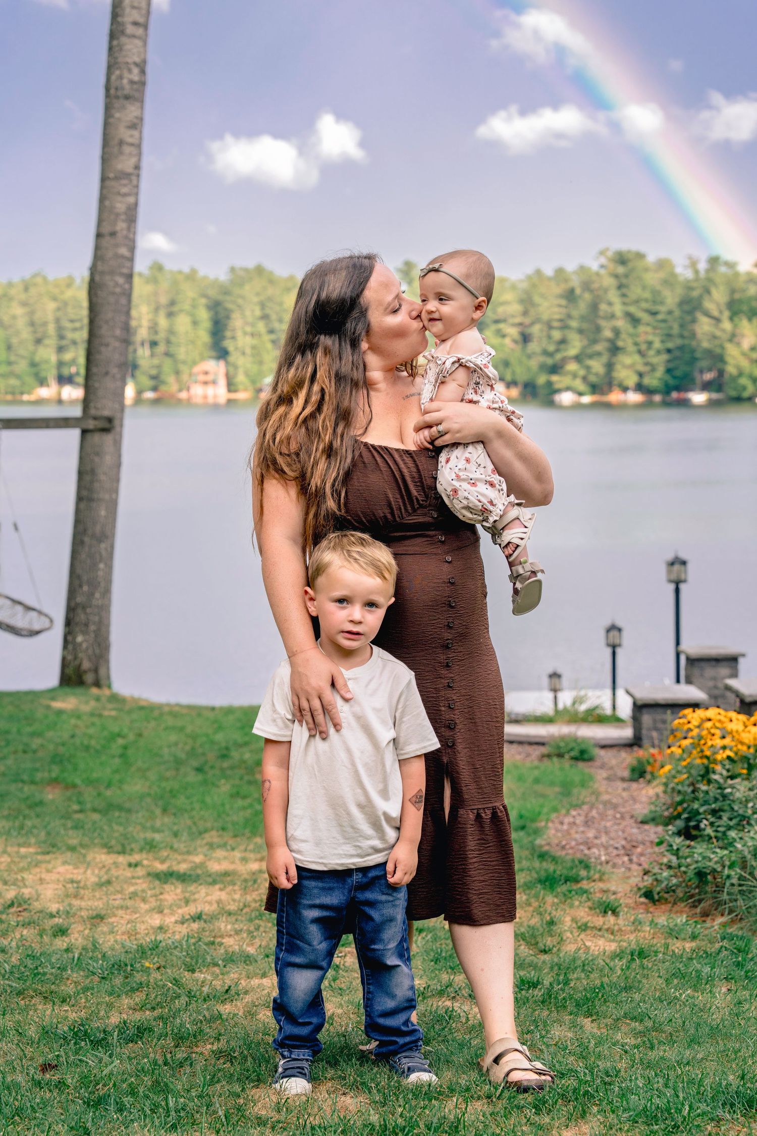 A family stands by a lakeside on a sunny day with a rainbow visible in the background and flowers in the foreground.