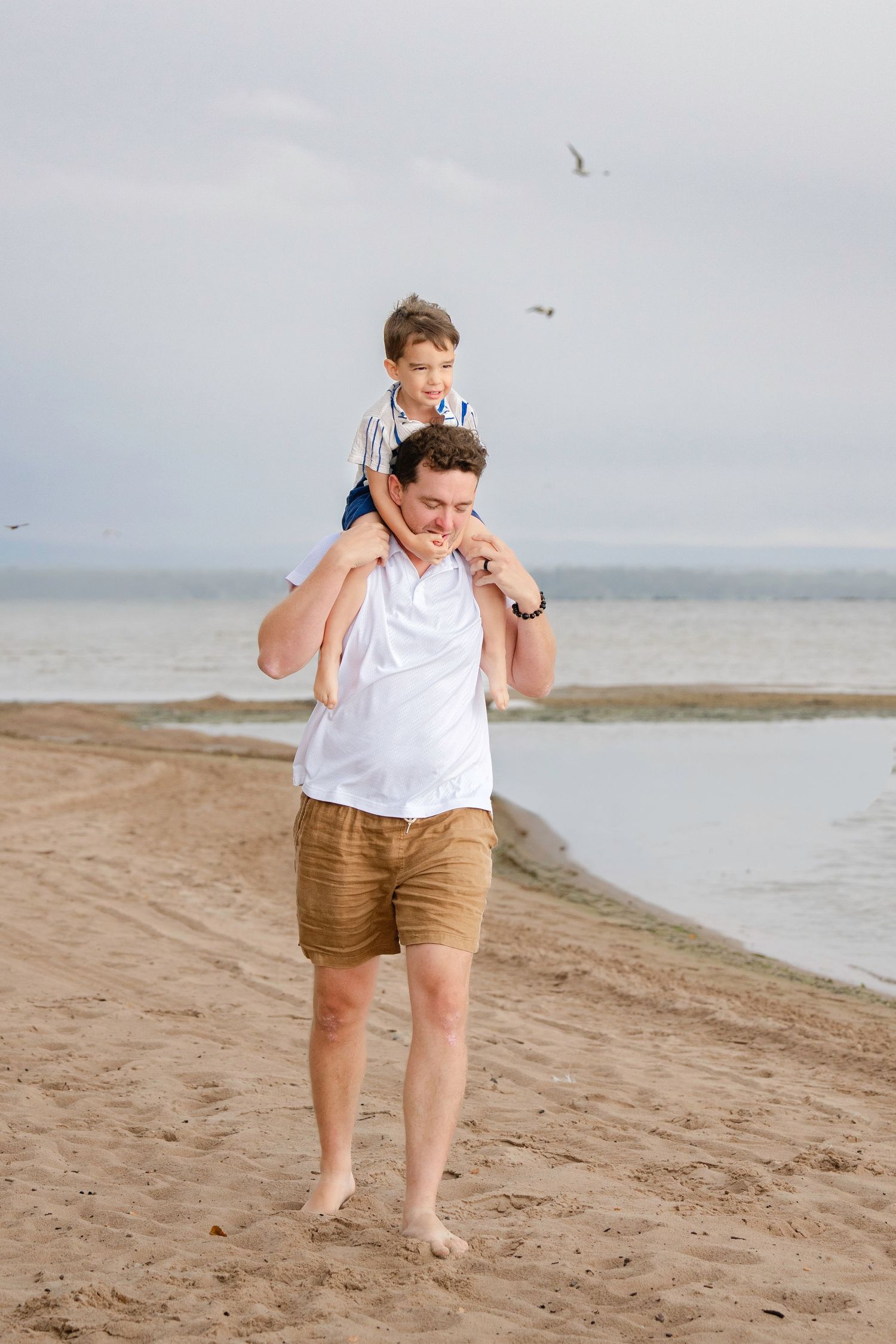 Person in white shirt and brown shorts walking and posing playfully along a sandy beach shoreline.