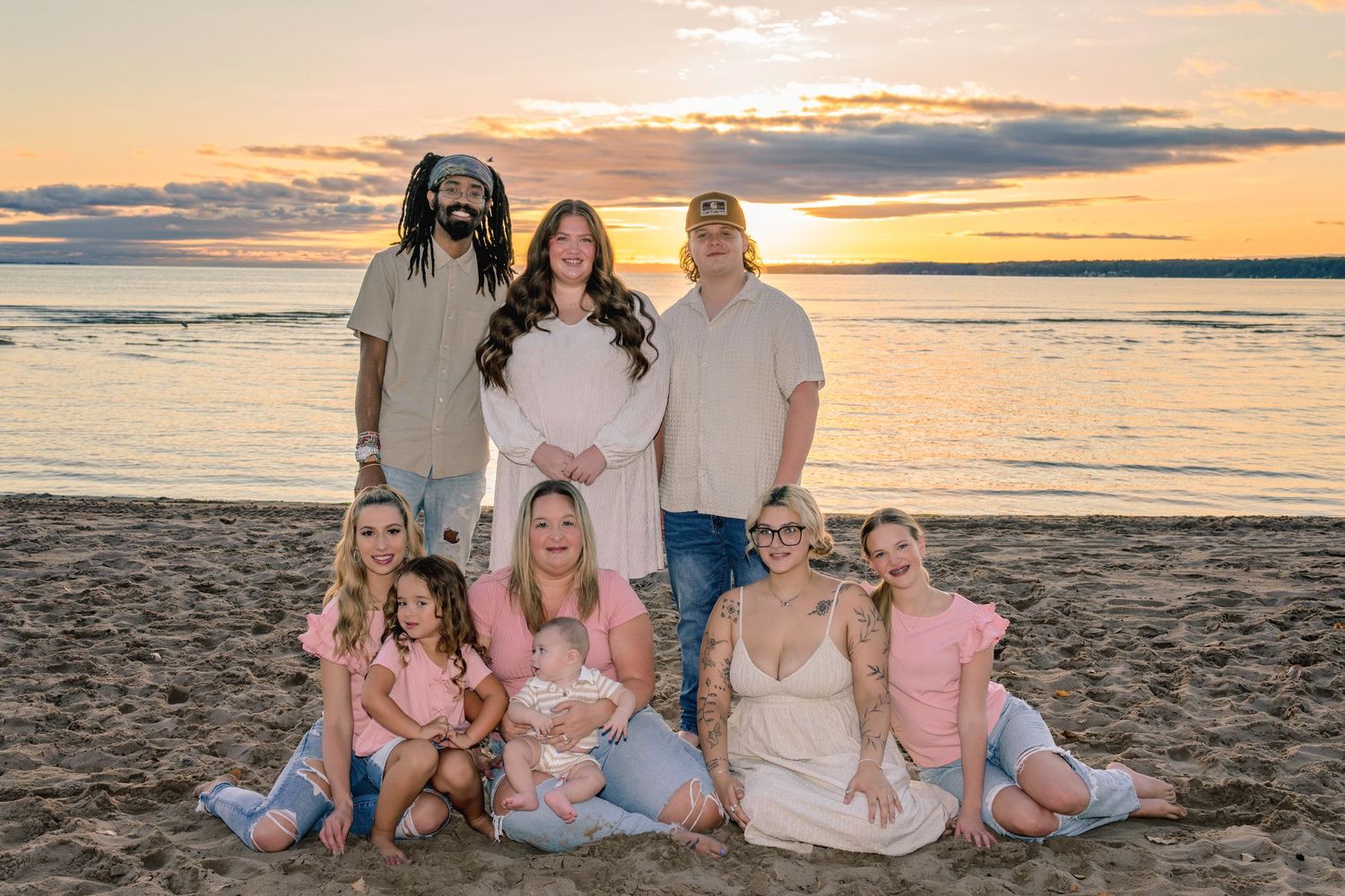Extended family group gathered on beach at sunset wearing coordinated pink and white outfits.