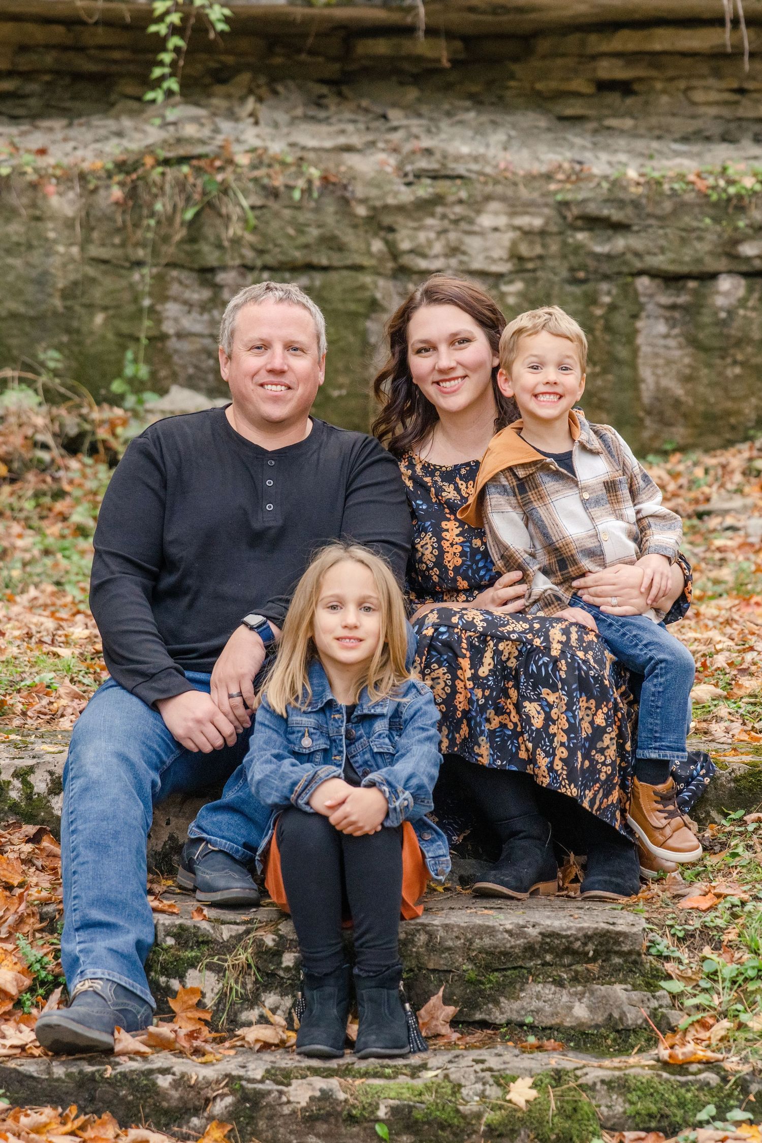 A family of four sits together on stone steps surrounded by autumn leaves and a rock wall backdrop.
