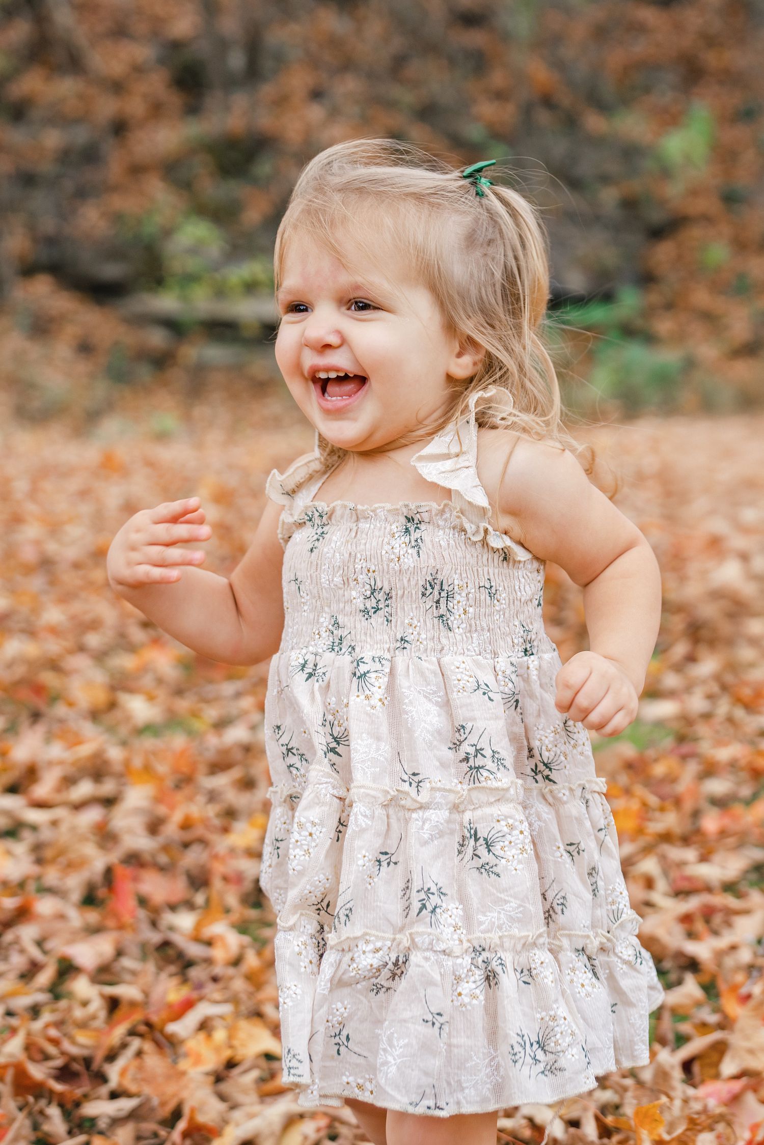 A child in a floral dress plays joyfully among fallen autumn leaves in an outdoor setting.