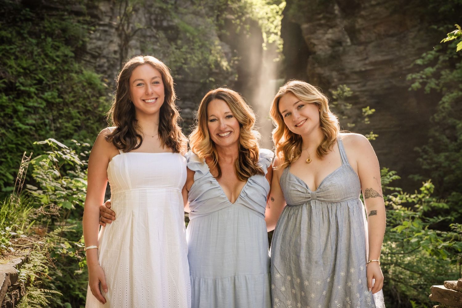 A group in coordinating light gray and white dresses poses together in a sunlit forest setting.