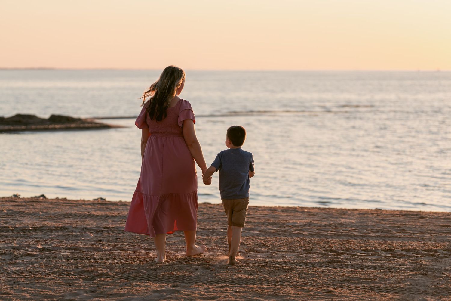 Mother and child silhouetted against sunset sky walking together on sandy beach by ocean.