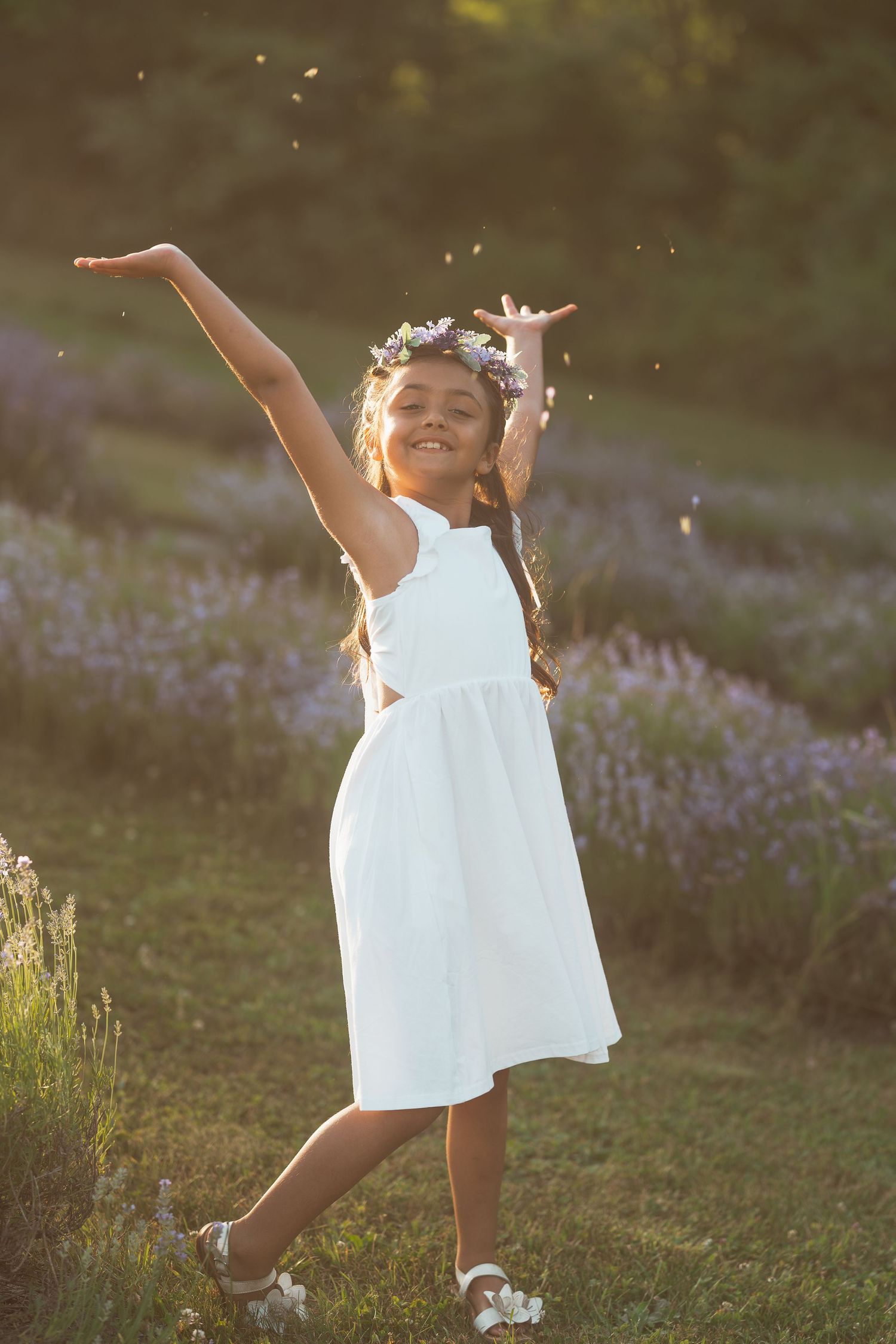 A child in a white dress twirls joyfully among purple wildflowers at golden hour. Family photos.