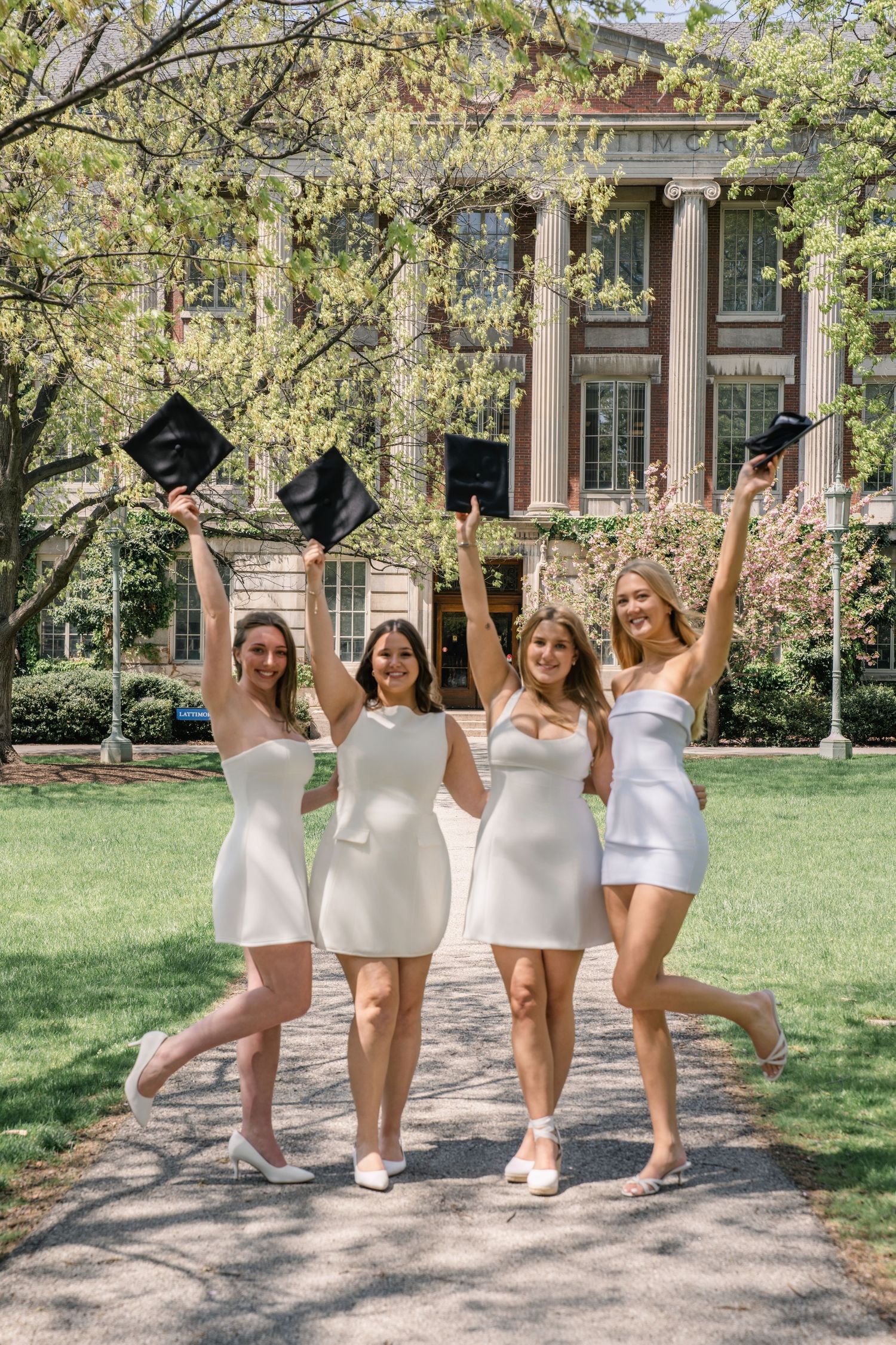 Four graduates in white dresses celebrate by tossing their caps on a sunny spring day at a university campus.