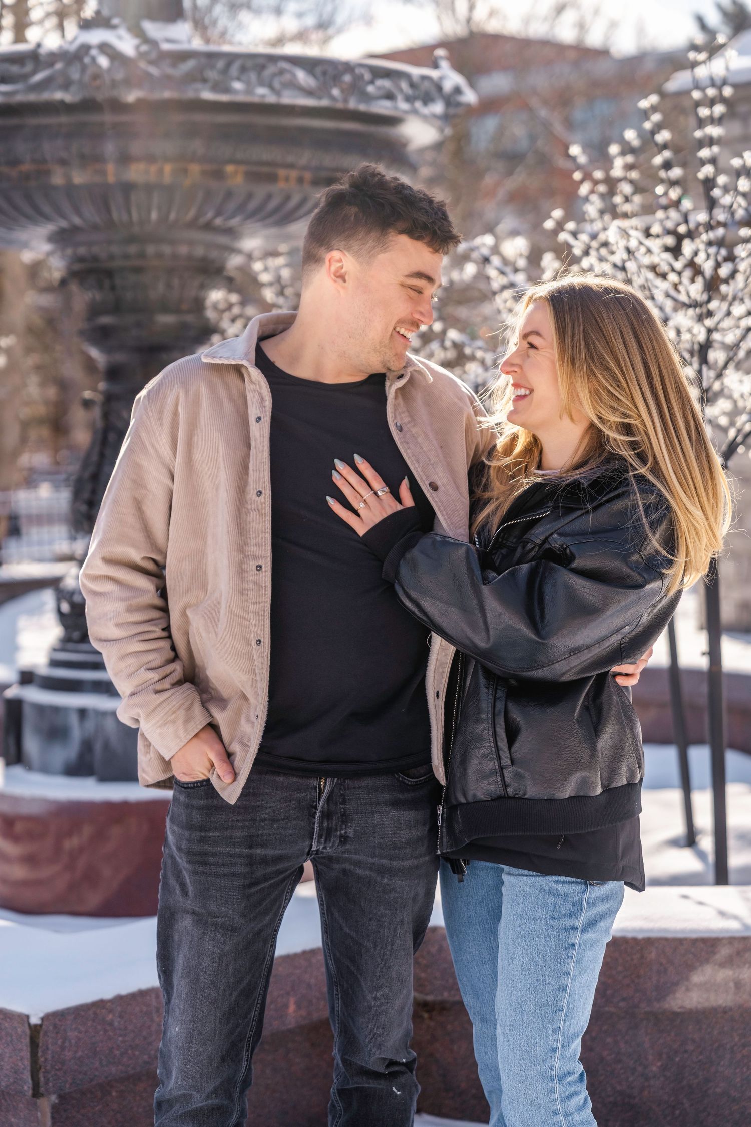 A happy couple smiles at each other outdoors in winter, the woman in a black jacket resting her hand on the man's chest.