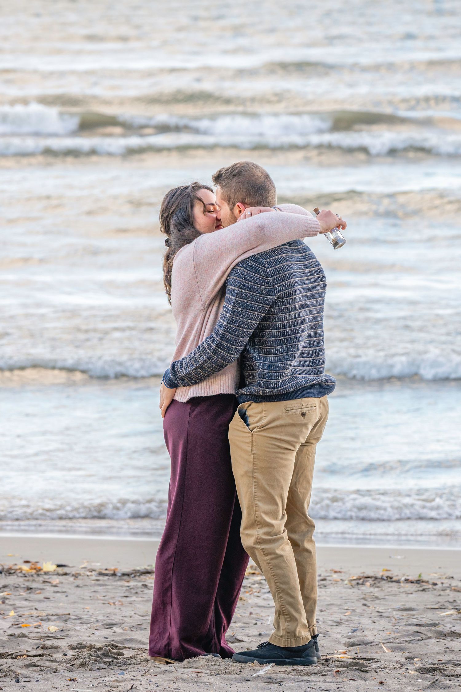 A romantic couple embraces while standing together on a beach with waves in the background.