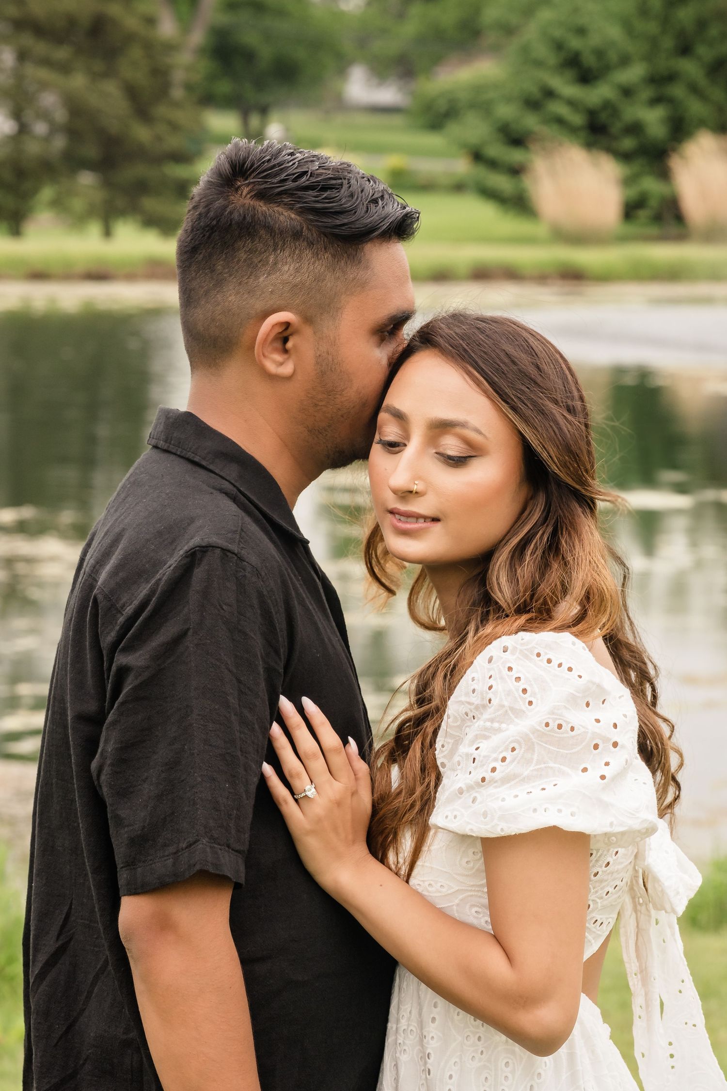 A romantic couple embraces near a serene lake with trees in the background on a sunny day.