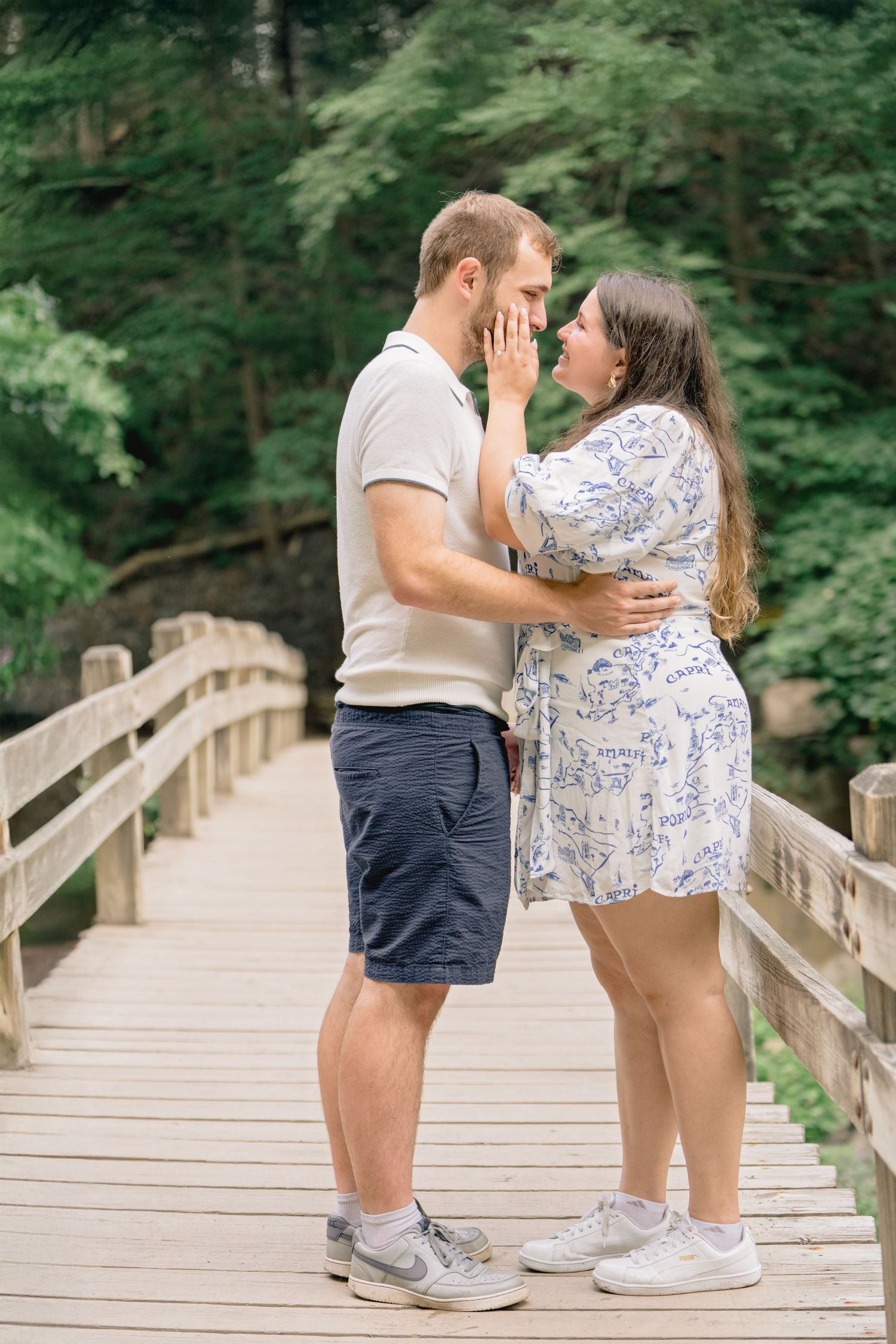 A couple shares a tender moment while standing on a wooden bridge surrounded by lush green forest.