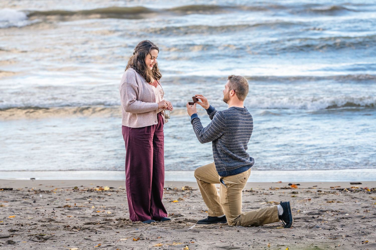 A couple shares a romantic beach proposal moment as waves crash in the background at sunset.