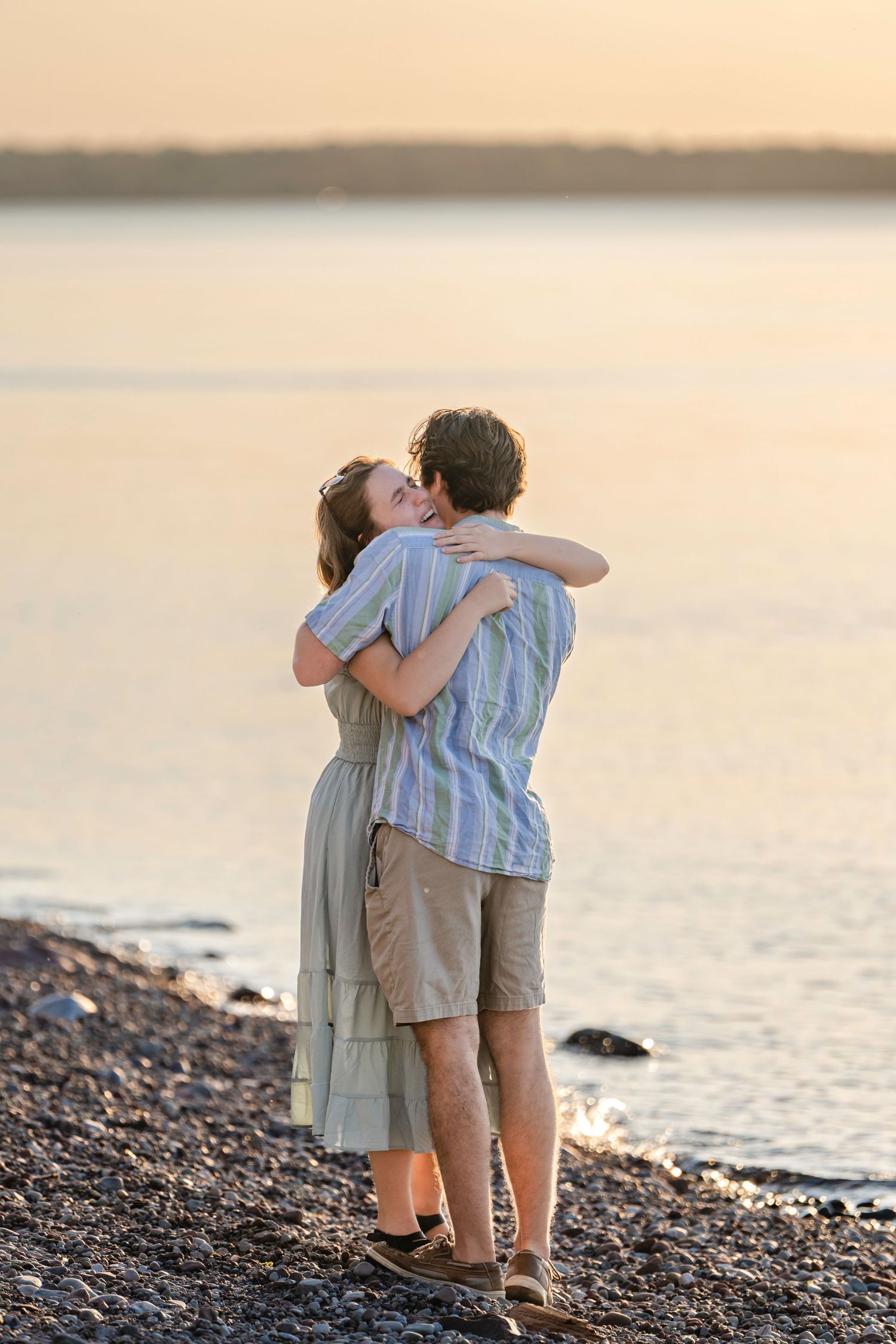 A romantic couple embraces while standing on a rocky beach at sunset with calm waters in the background.