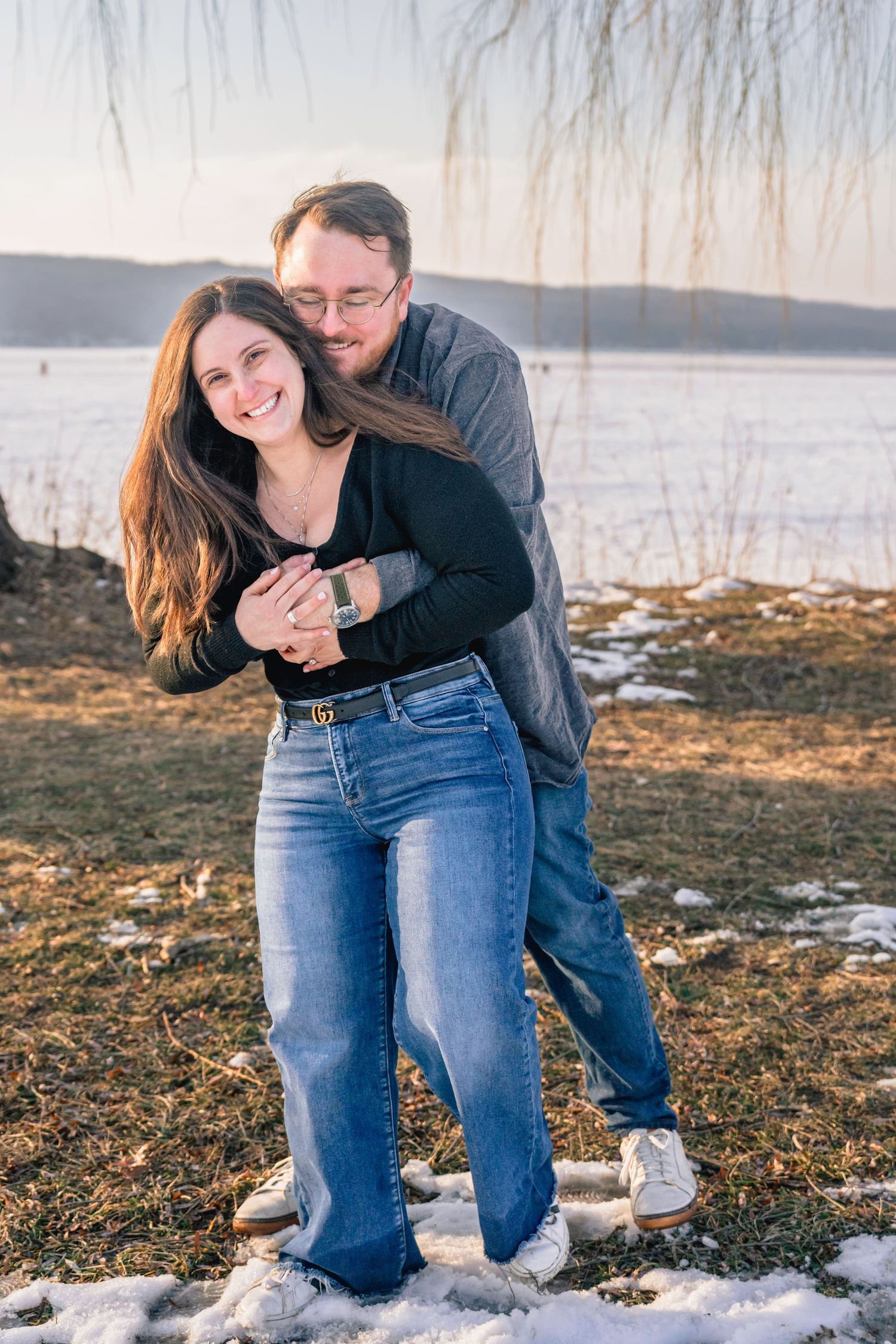 Couple embracing outdoors in winter, smiling by a frozen lake with snow on the ground and willow tree.