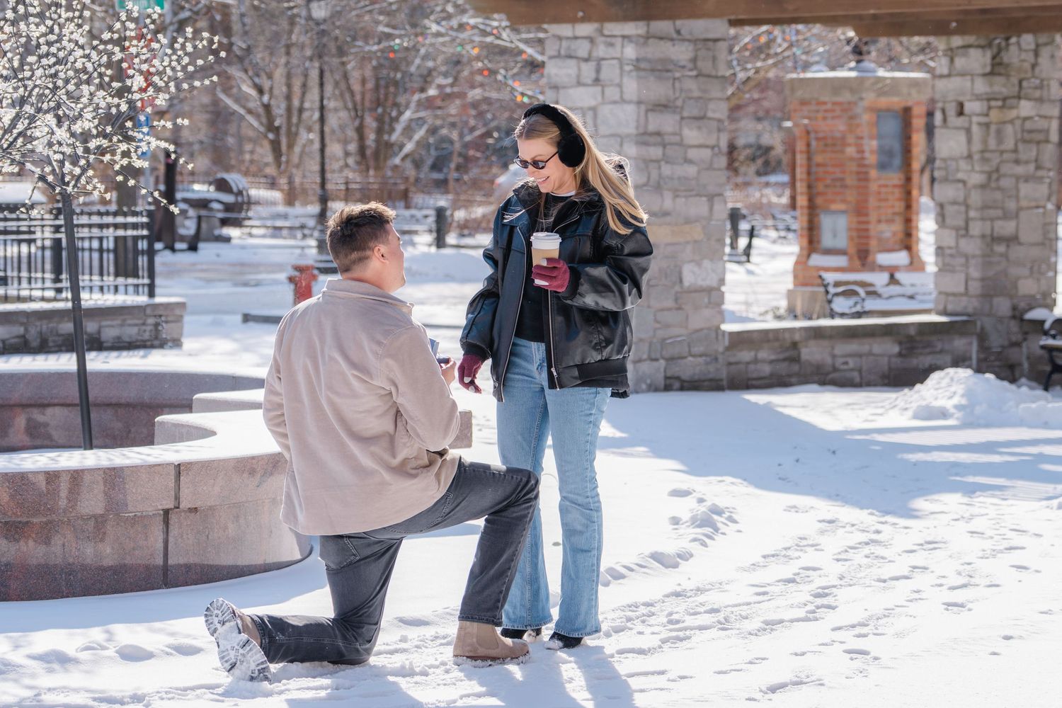 Man proposing on one knee to woman in snowy outdoor plaza on a sunny winter day.