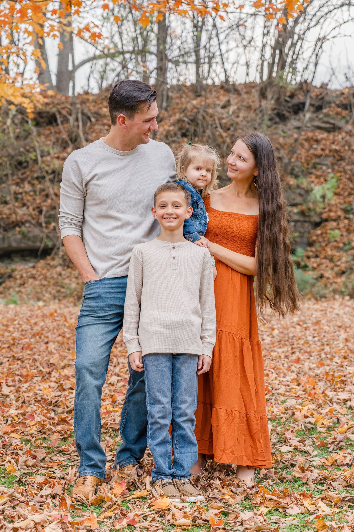 A family poses together on fallen autumn leaves in the woods wearing coordinated neutral and orange outfits.