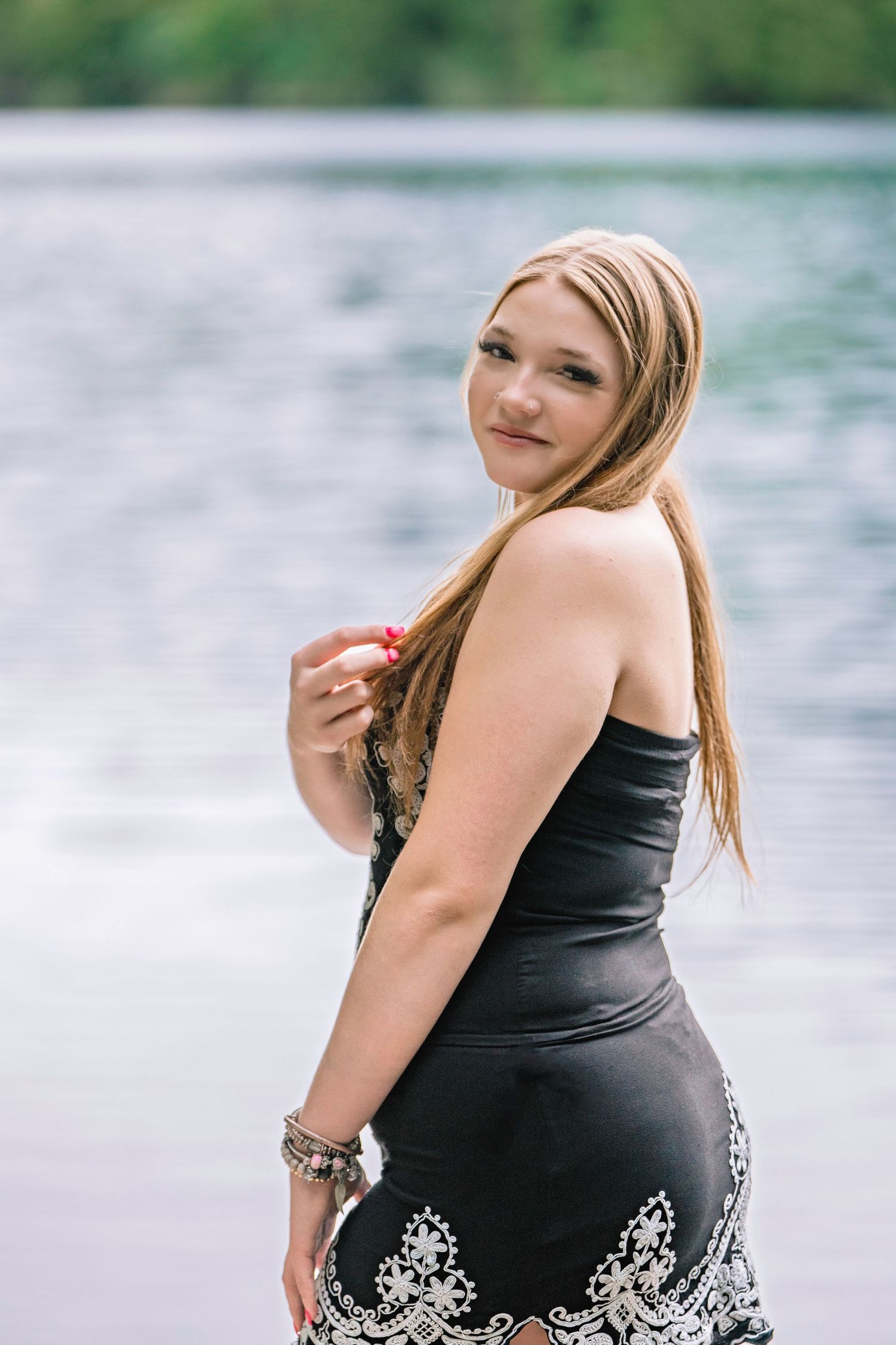 Person in black dress with decorative details poses near waterfront with blurred lake background.
