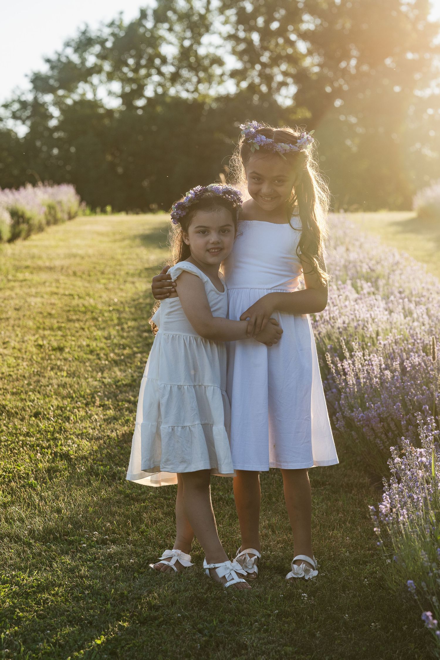 Two sisters in white dresses stand together in a sunlit field of lavender at golden hour.