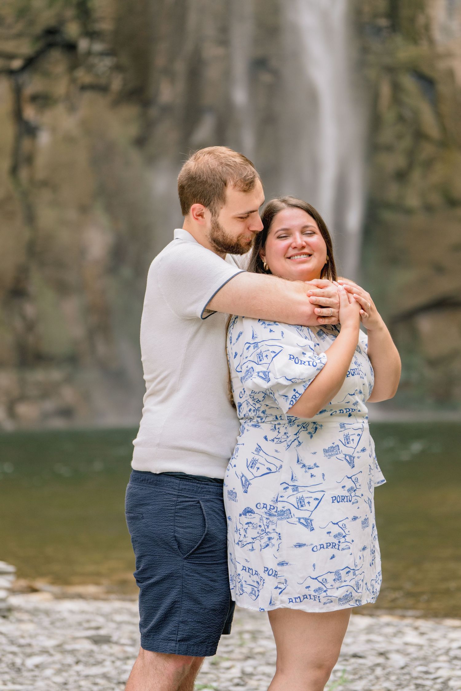 A couple shares a joyful embrace by a waterfall, with the woman wearing a blue and white patterned dress.