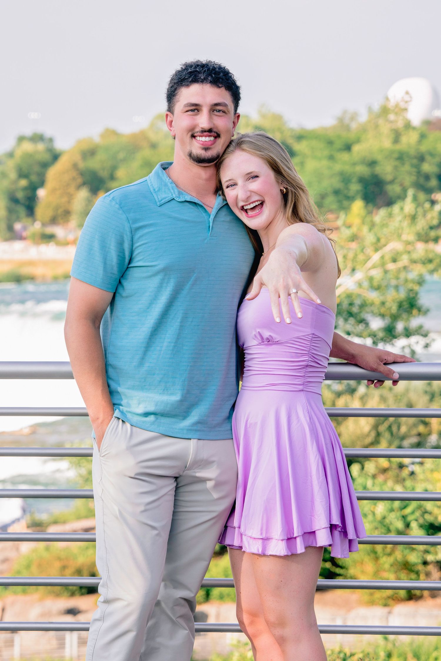 A couple embraces on an outdoor viewing platform with trees and water in the background.