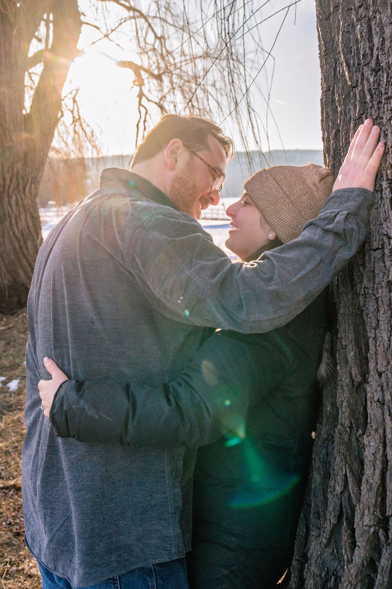 Couple sharing a romantic moment leaning against a tree outdoors in winter sunlight.