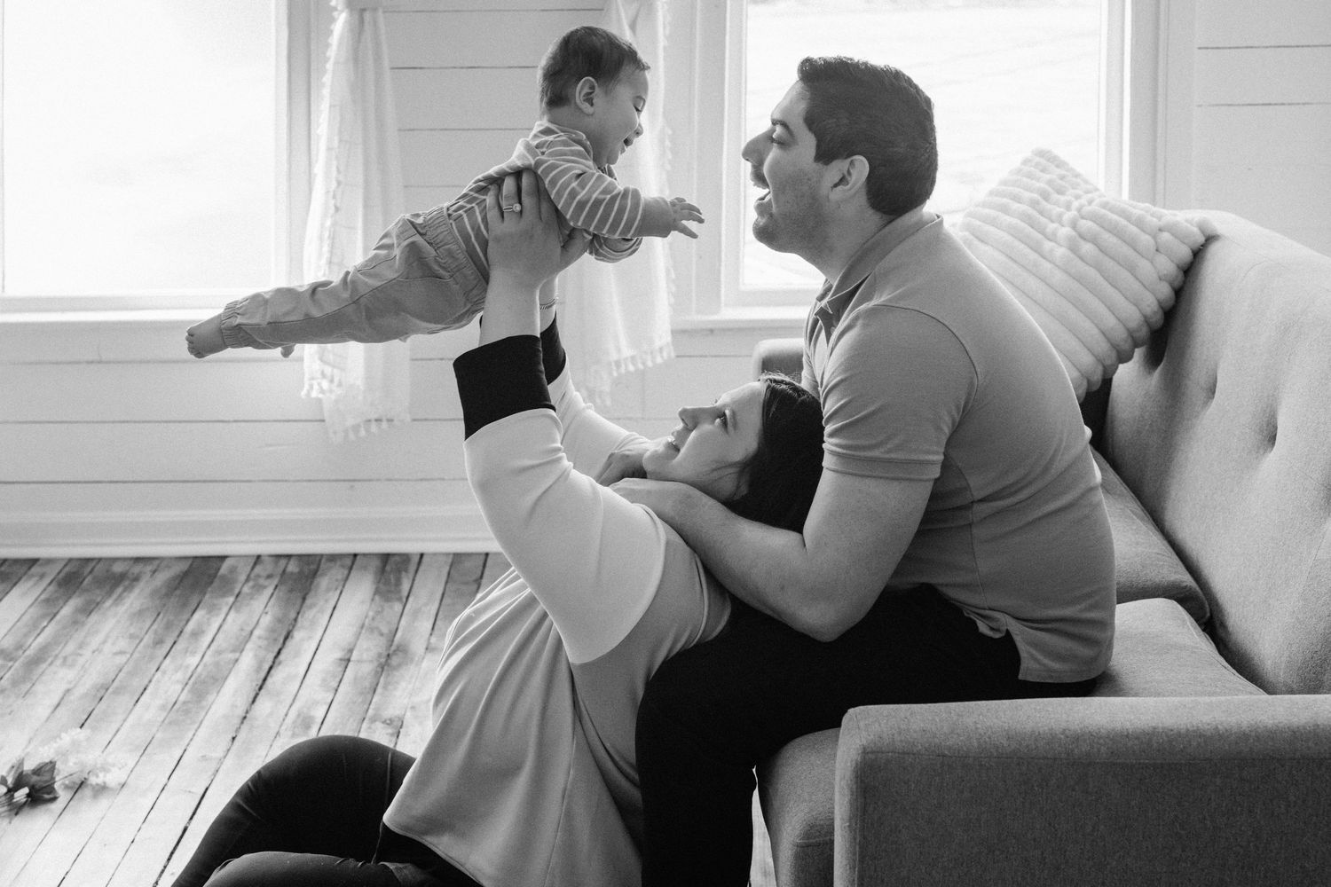 Black and white photo of parents playing with baby, father lifting child while mother reclines on living room floor.