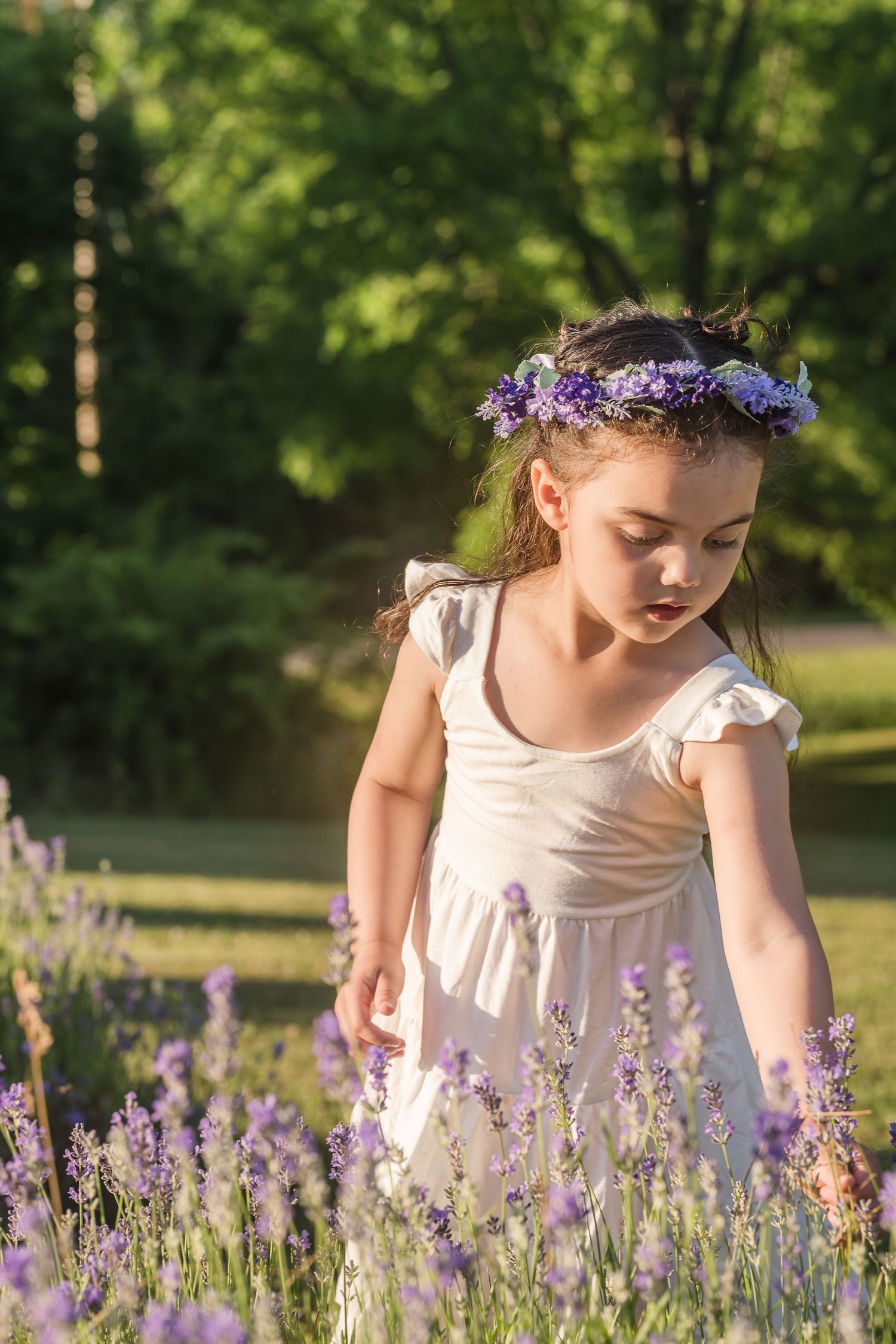 A person wearing a white dress leans over to smell blooming purple lavender flowers in a sunny garden setting.