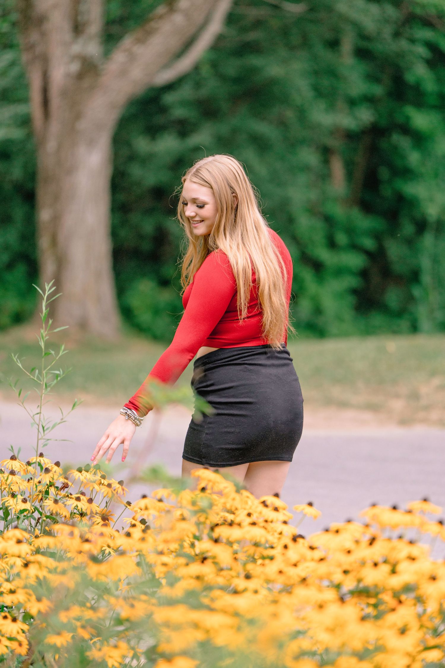 Woman in red top and black skirt bending down to admire yellow wildflowers in a garden path.