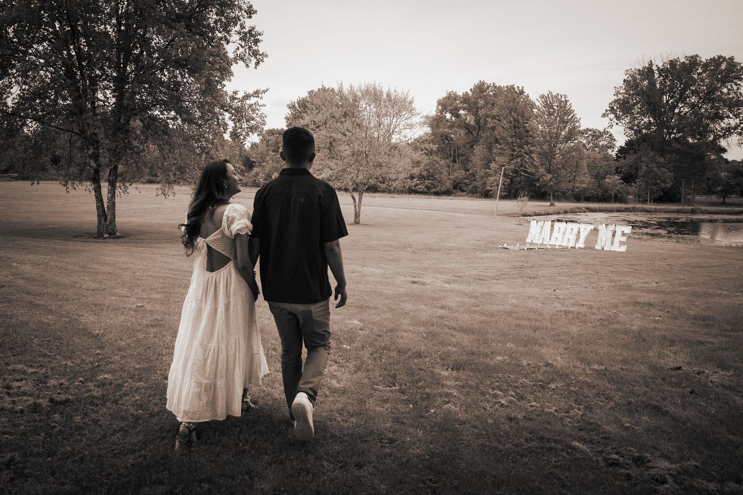 A couple walks away from the camera across an autumn field with a 'Marry Me' sign visible in the distance.