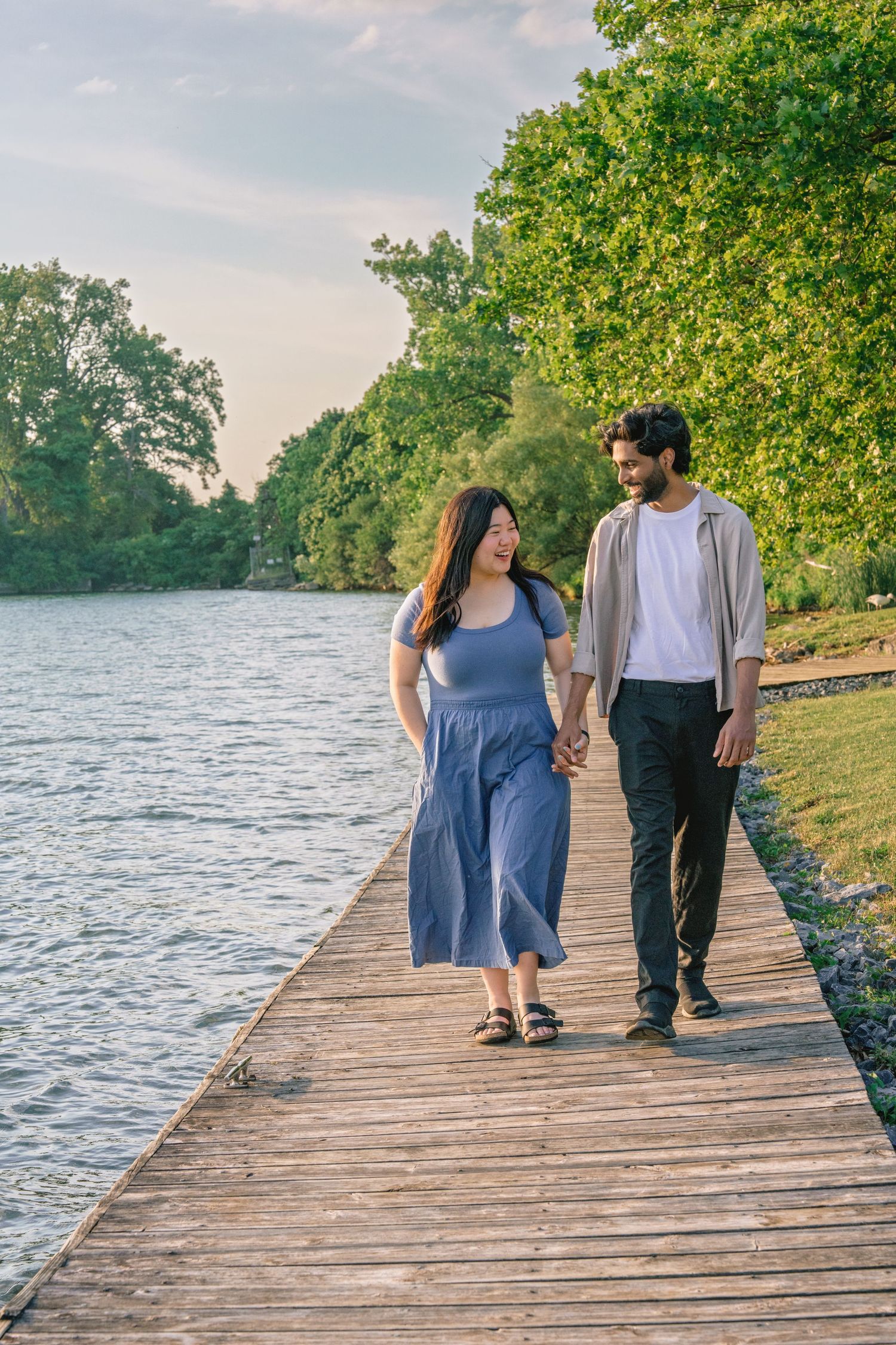 A couple walks together on a wooden boardwalk by a serene lake surrounded by lush green trees on a summer day.