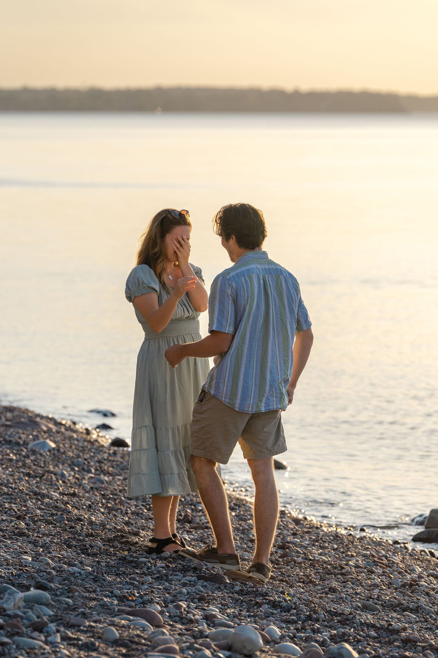 A couple shares a romantic moment on a rocky beach at sunset, sitting close together by the water.