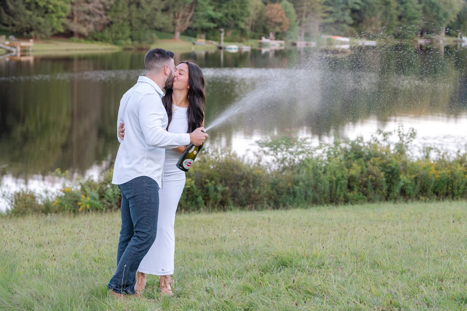 A couple celebrates with champagne by a lake during a special outdoor moment.