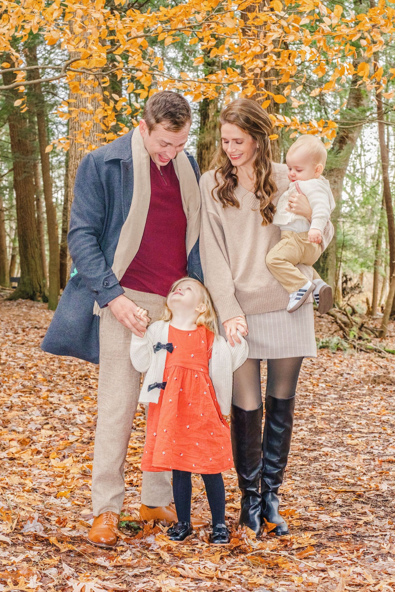 A family poses together on fallen autumn leaves under golden trees wearing coordinated neutral-toned outfits.