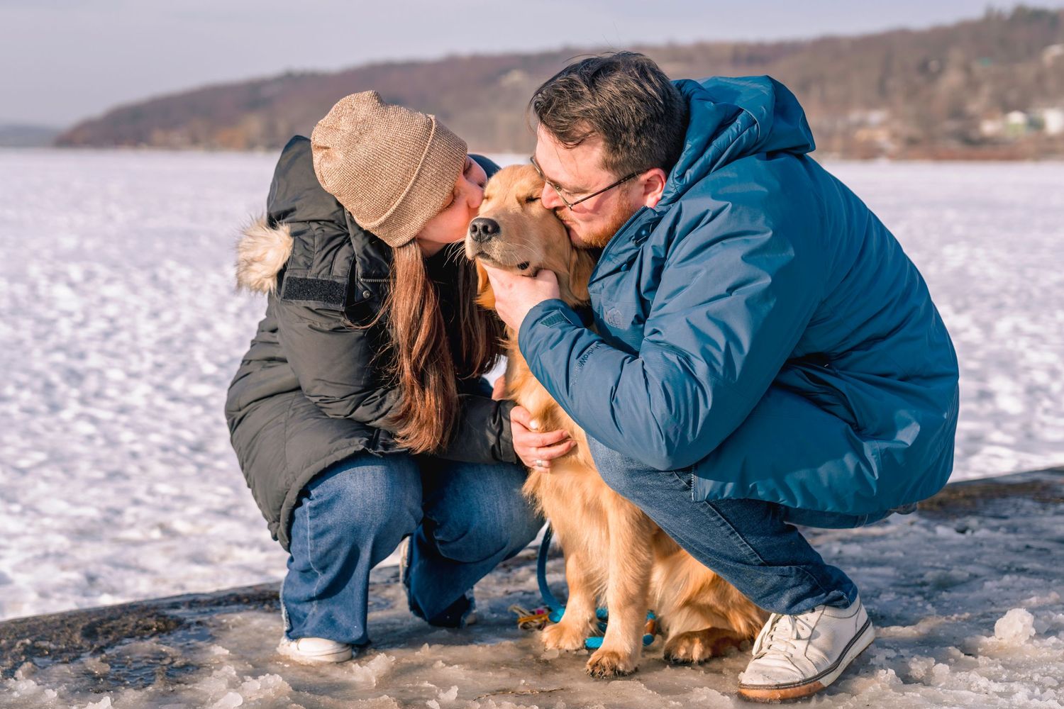 Couple crouching in snow kissing a golden retriever dog outdoors in winter.