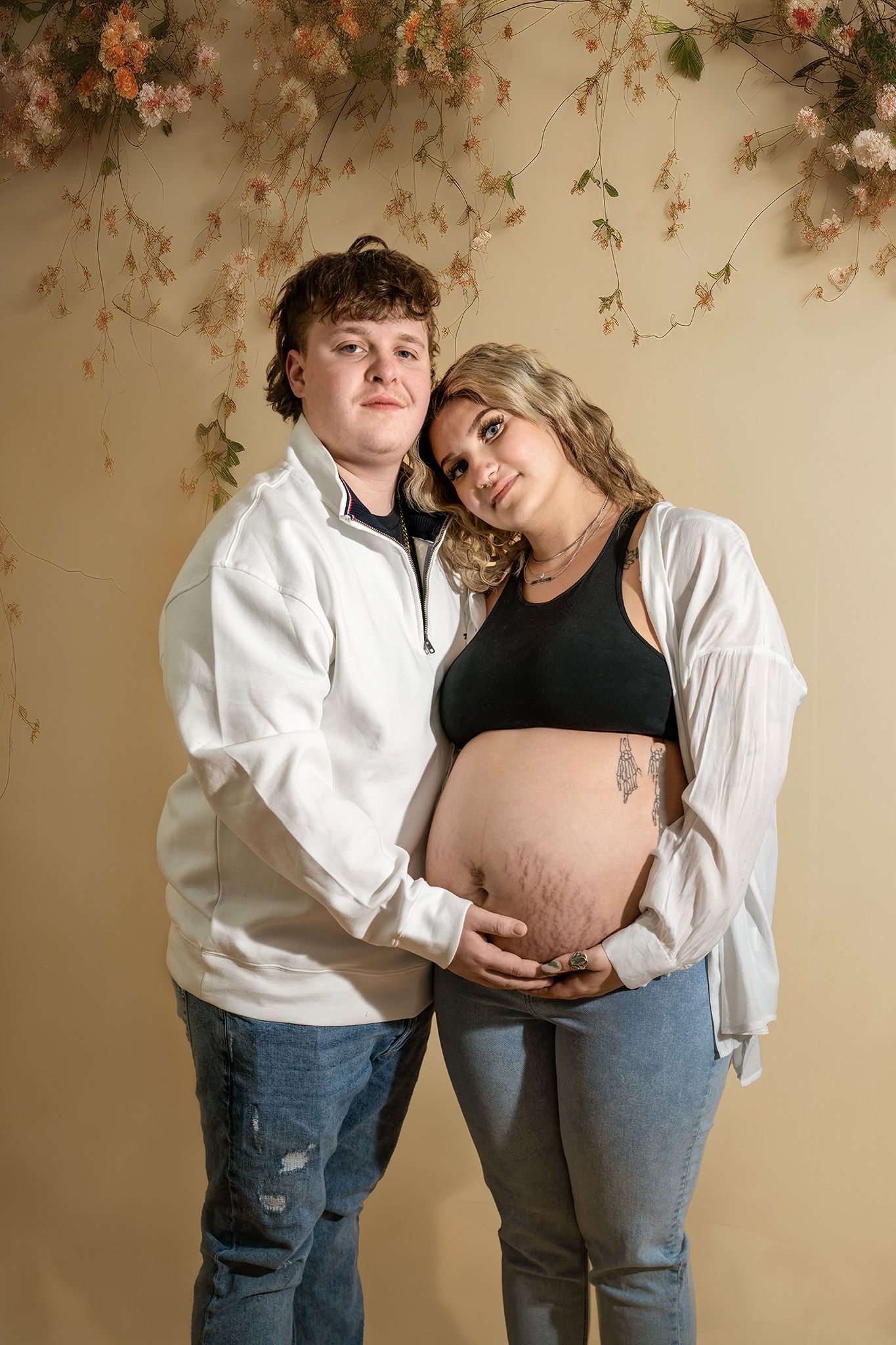 A couple in casual clothing poses for a maternity photo against a floral backdrop with dried flowers.