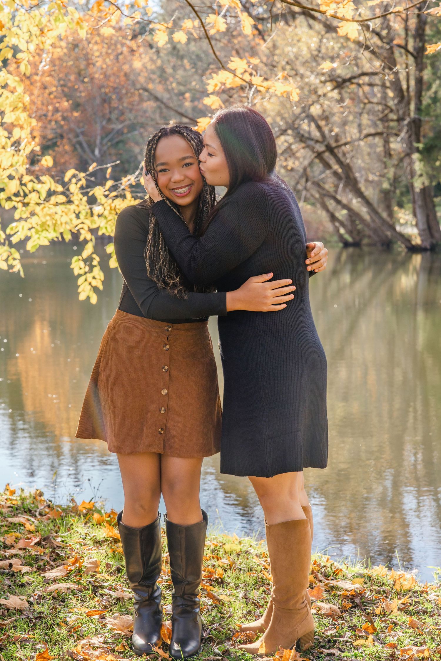 Two people share an embrace by a lake surrounded by autumn foliage and fallen leaves on a peaceful afternoon.