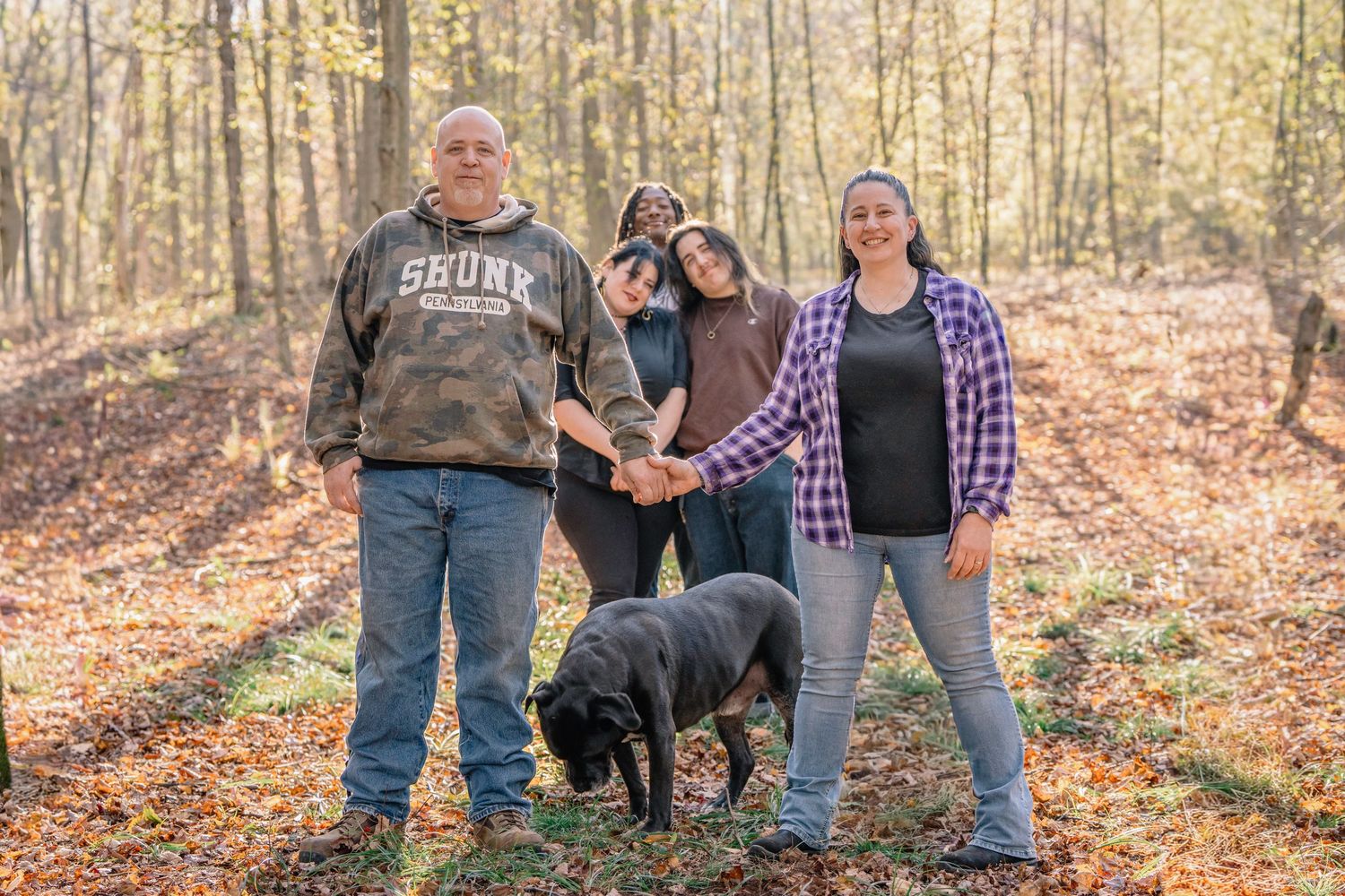 A group stands together with their black dog on fallen autumn leaves in a sunlit forest.