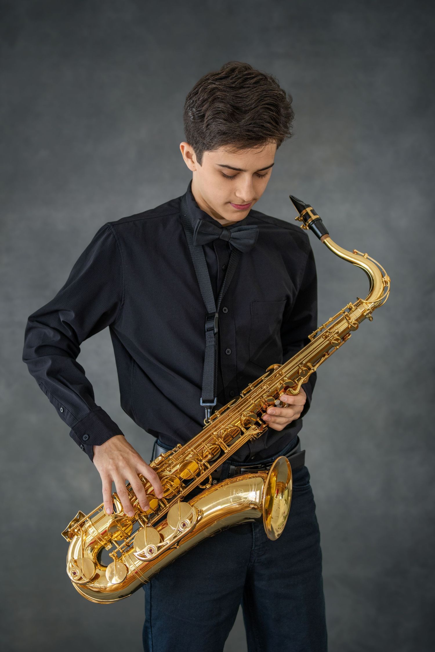 Young musician in black shirt plays a shiny brass saxophone against a dark studio backdrop. Senior photography