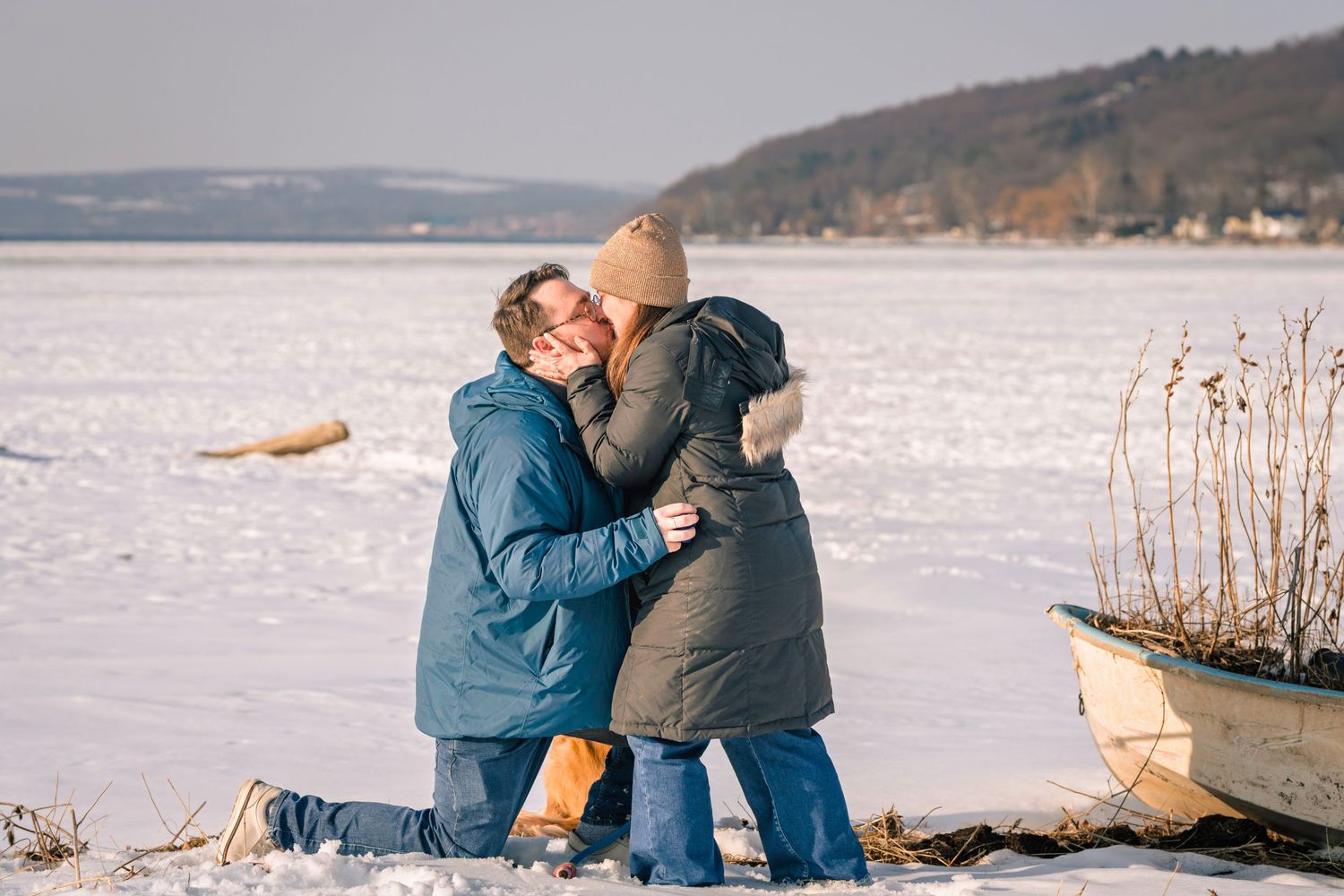 Couple sharing a romantic kiss on a snowy frozen lake shore in winter, wearing warm jackets outdoors.