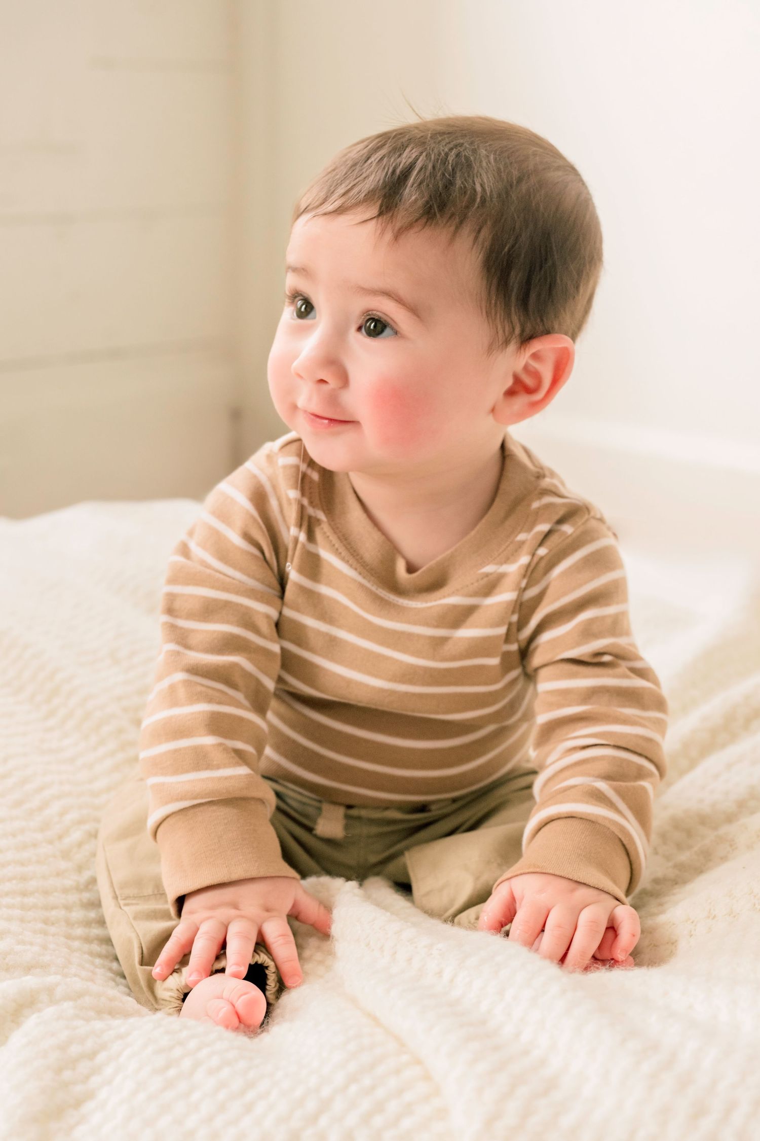 Smiling baby in striped tan onesie sitting on white blanket.