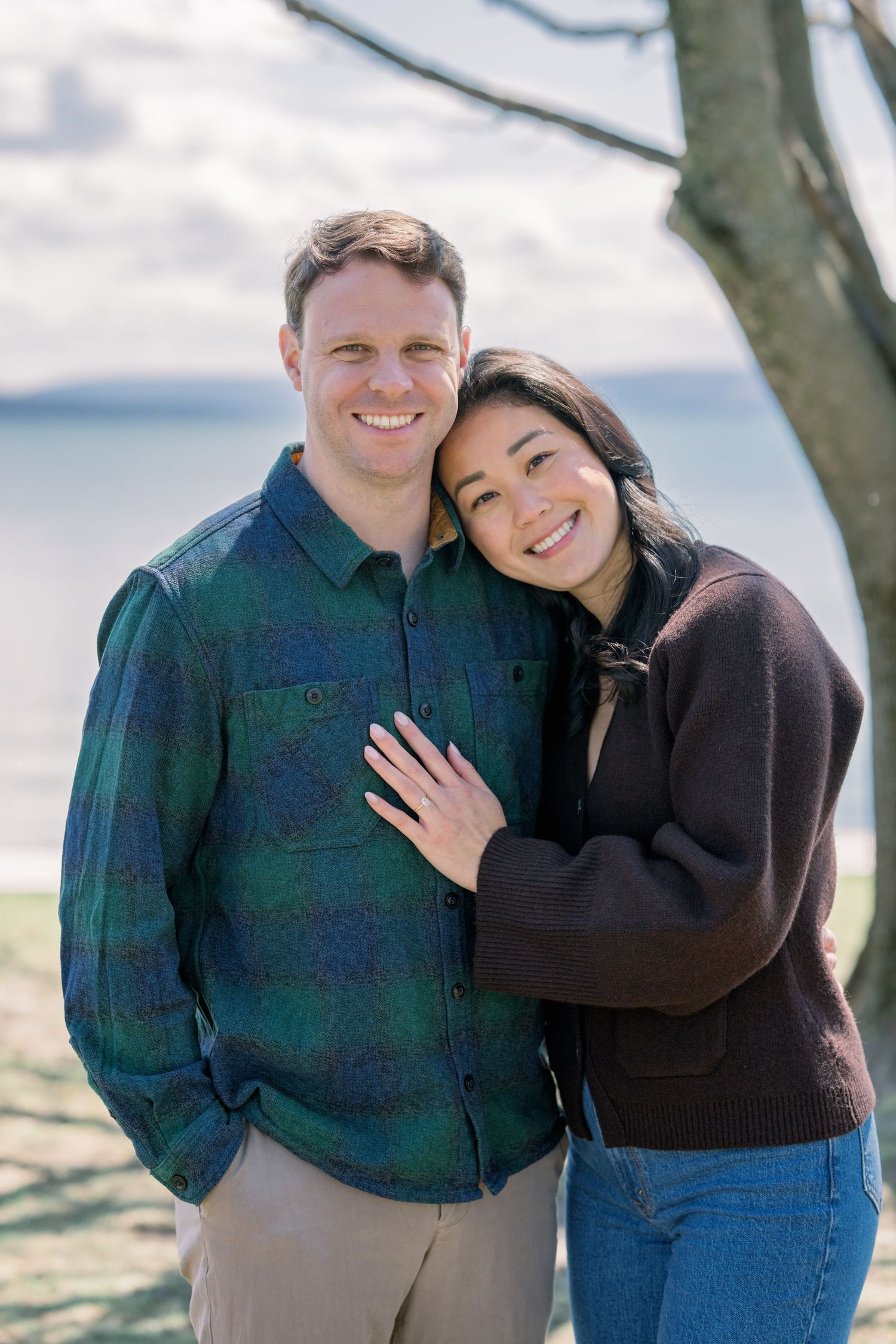 A couple poses together outdoors near a lake on a sunny day, with one wearing a plaid shirt and the other in a dark sweater.