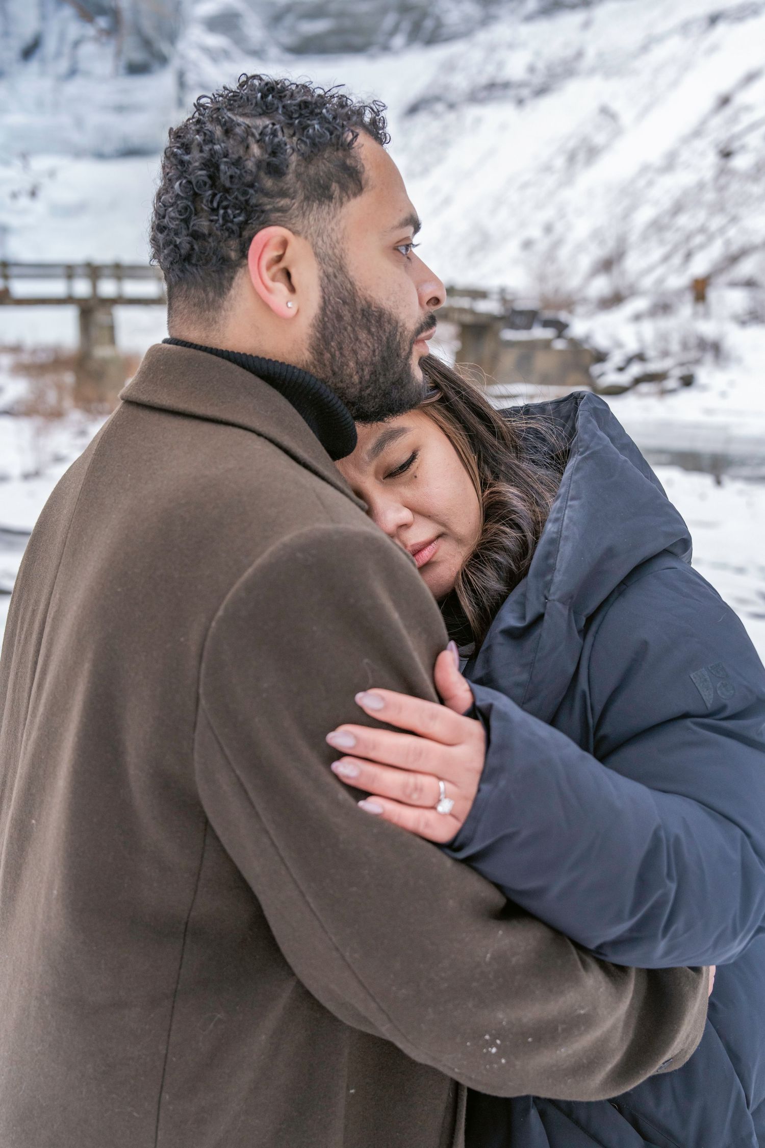 Two people hold each other close while standing in a snow-covered landscape during winter.