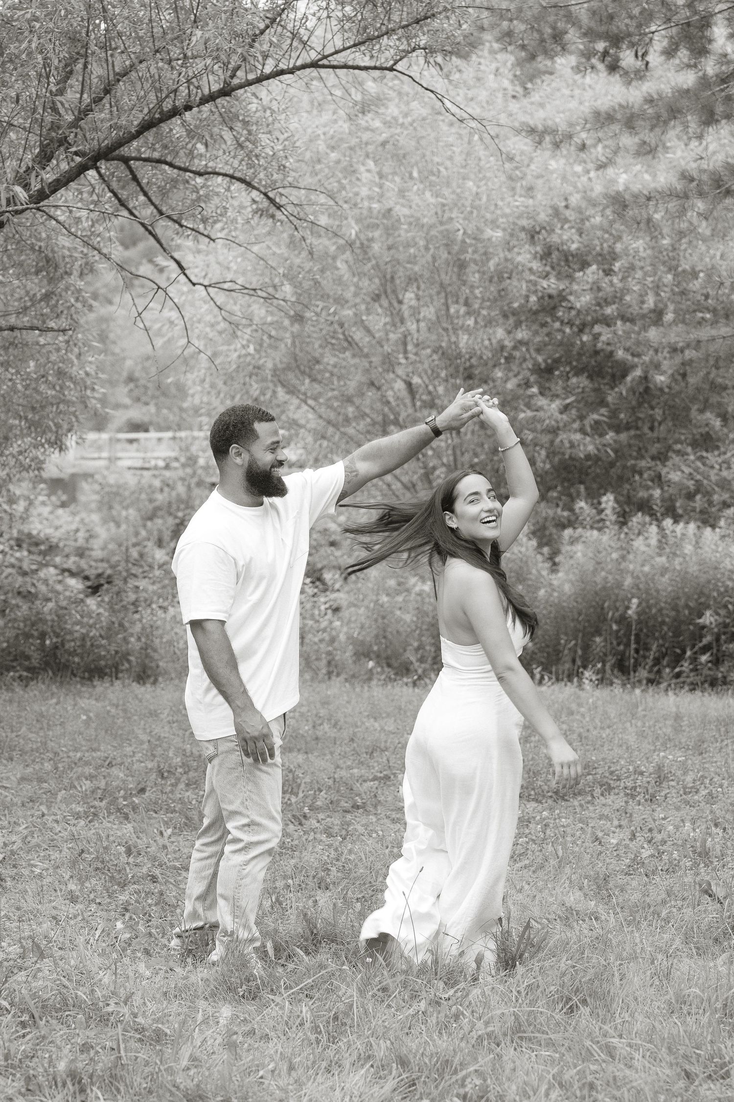 A black and white photo captures a couple dancing together in a meadow with natural scenery.