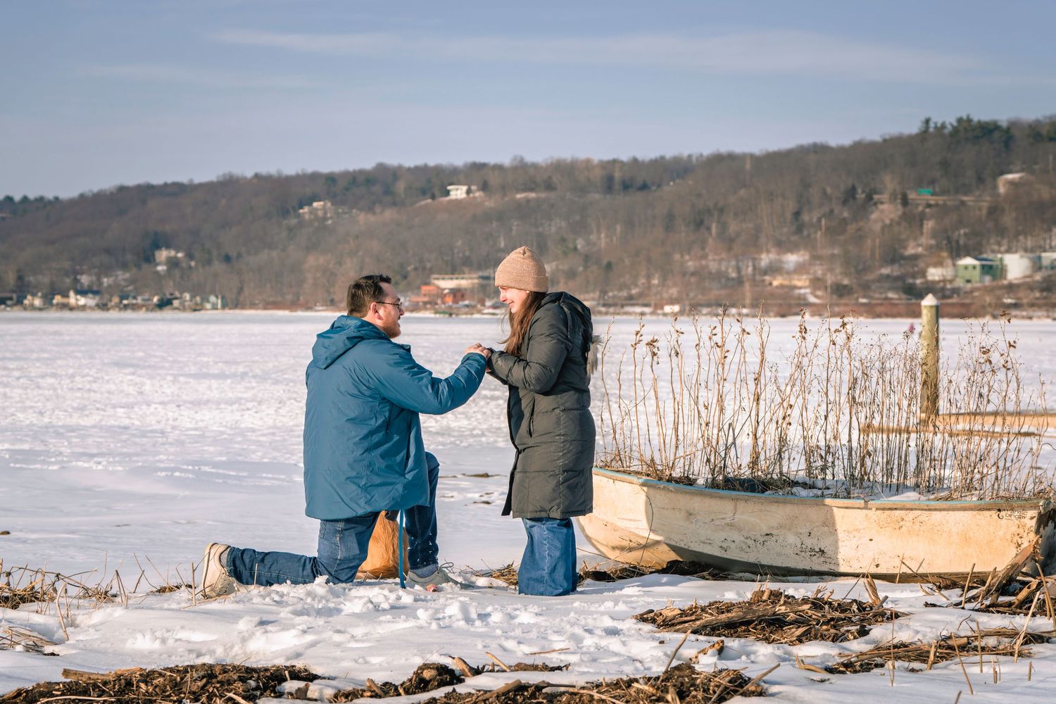 Man proposing to woman on snowy lakeside with vintage rowboat, romantic winter marriage proposal outdoors.