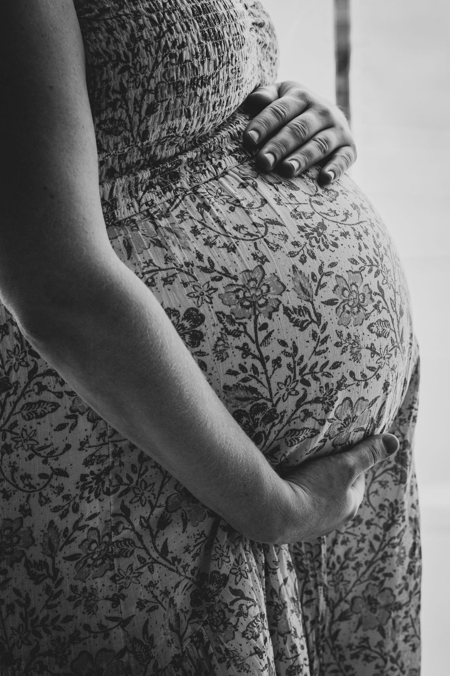 Black and white close-up of pregnant belly in a floral dress with gentle hands cradling the bump.