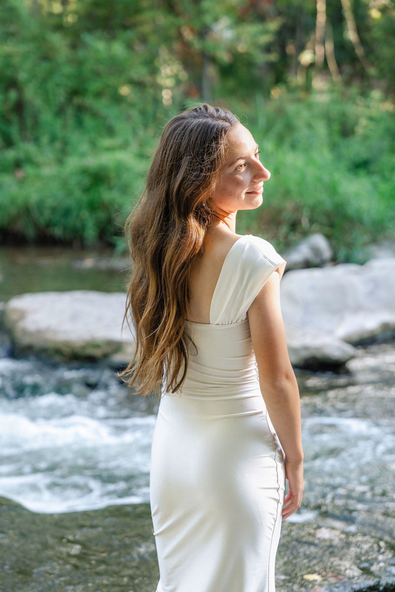Woman in white dress poses gracefully by rushing river waters in natural outdoor setting.