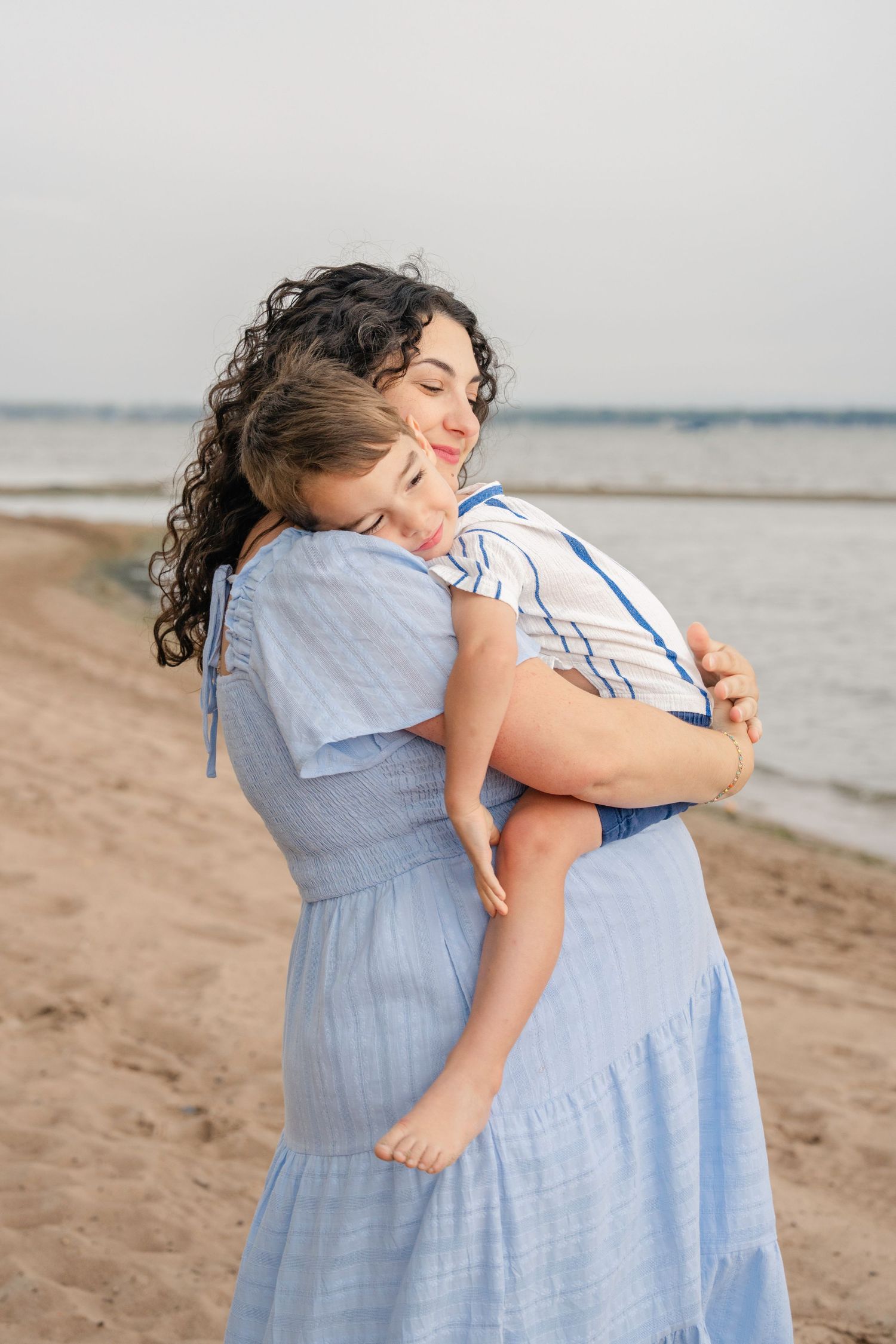 A series of tender embrace photos on a sandy beach with light blue dresses against a serene coastal backdrop.