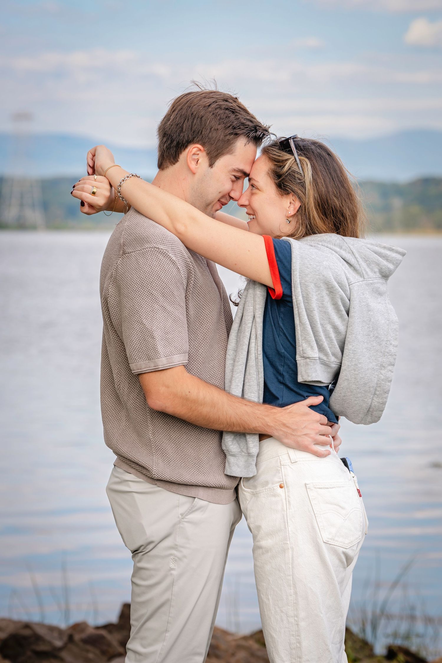 A couple embraces on a beach with water and mountains visible in the background.