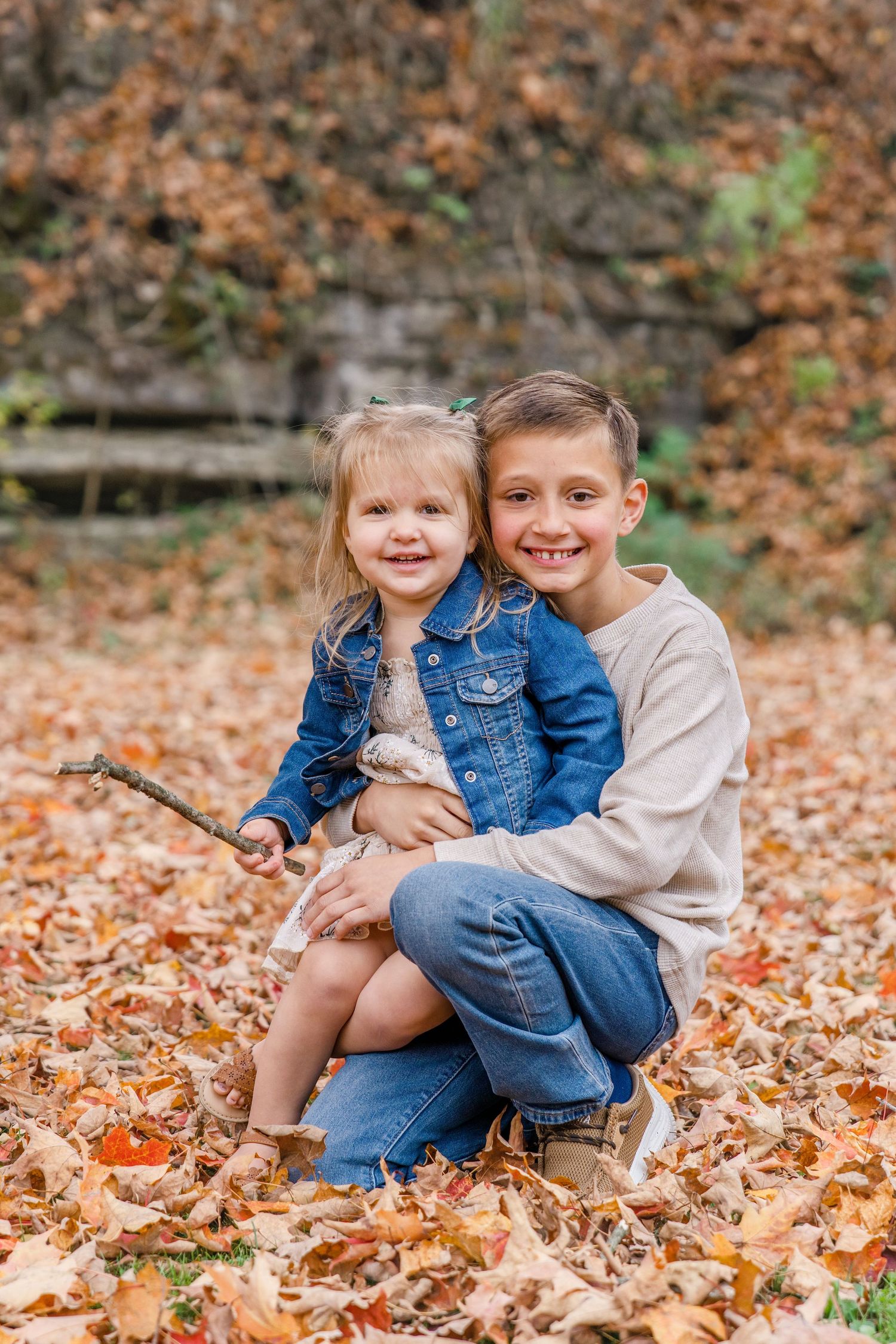 Two young children sit together on fallen autumn leaves in a forest setting.